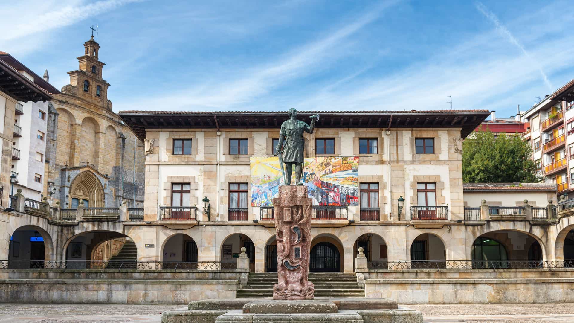 A statue of a Basque hero in the town square of Guernica, Spain, with the historic Casa de Juntas de Guernica building and a beautiful stone church in the background.