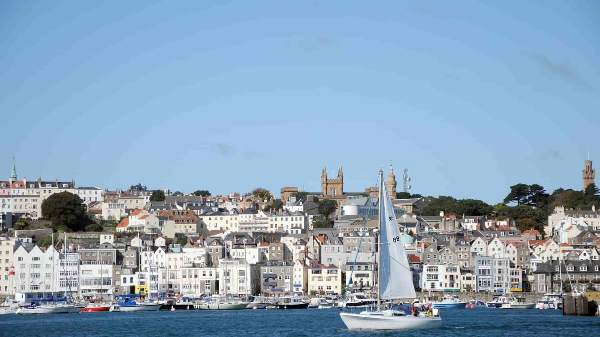 A sailboat gracefully sailing in the harbor of St. Peter Port, Guernsey, with the town's historic buildings and spires sprawling up the hillside in the background.