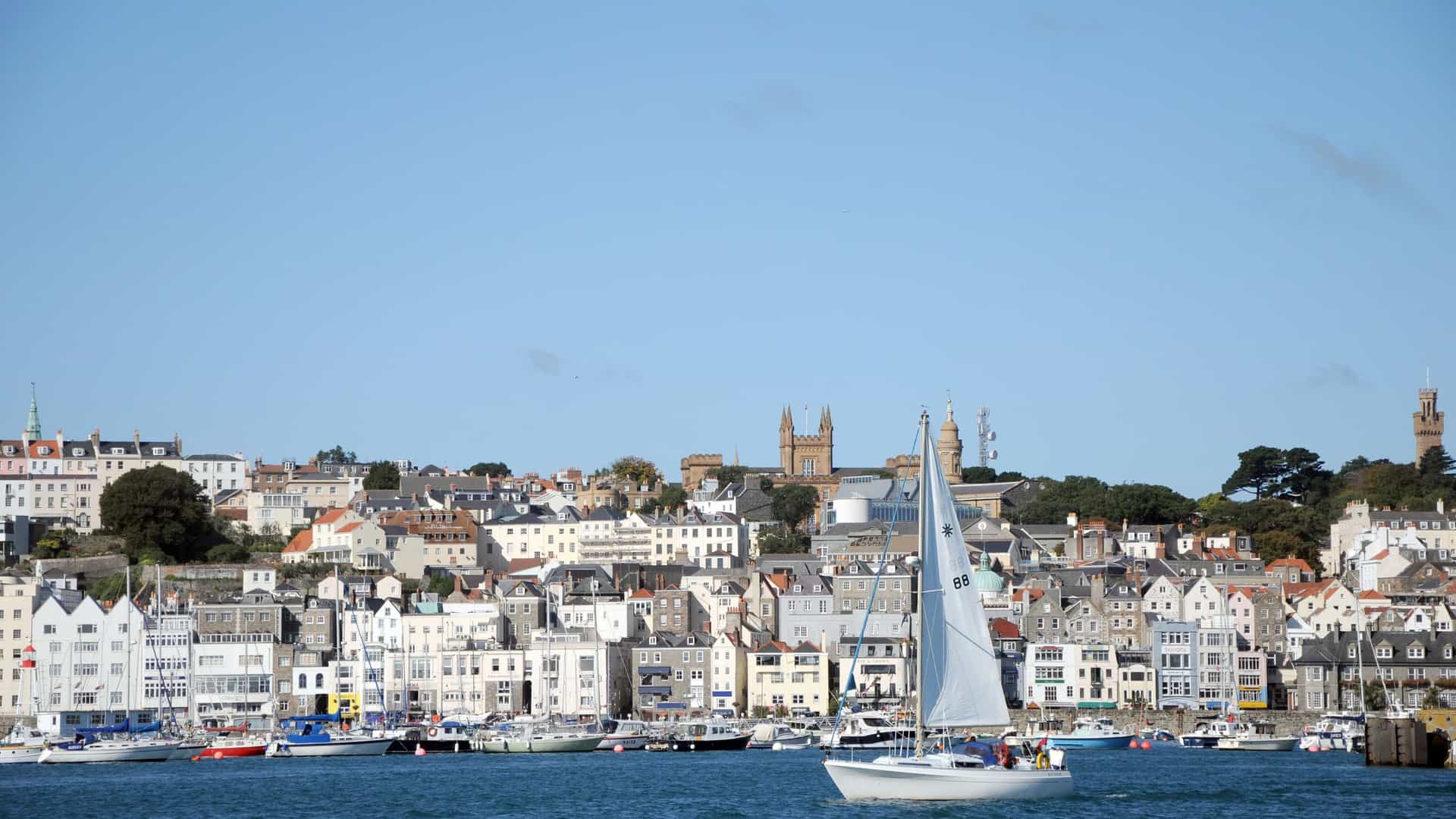 A sailboat gracefully sailing in the harbor of St. Peter Port, Guernsey, with the town's historic buildings and spires sprawling up the hillside in the background.