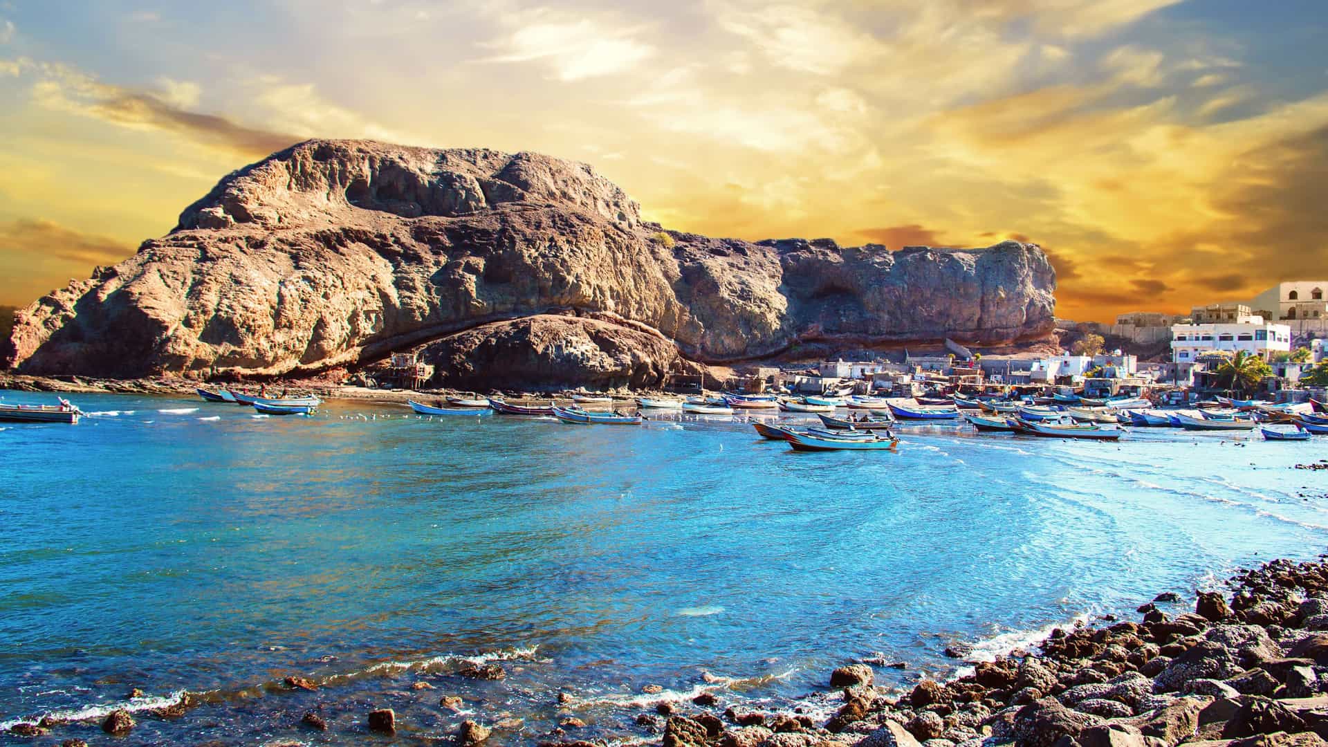 A vibrant sunset over the Gulf of Aden, with traditional fishing boats in the harbor near Aden, Yemen, with dramatic, rocky cliffs towering over the town.