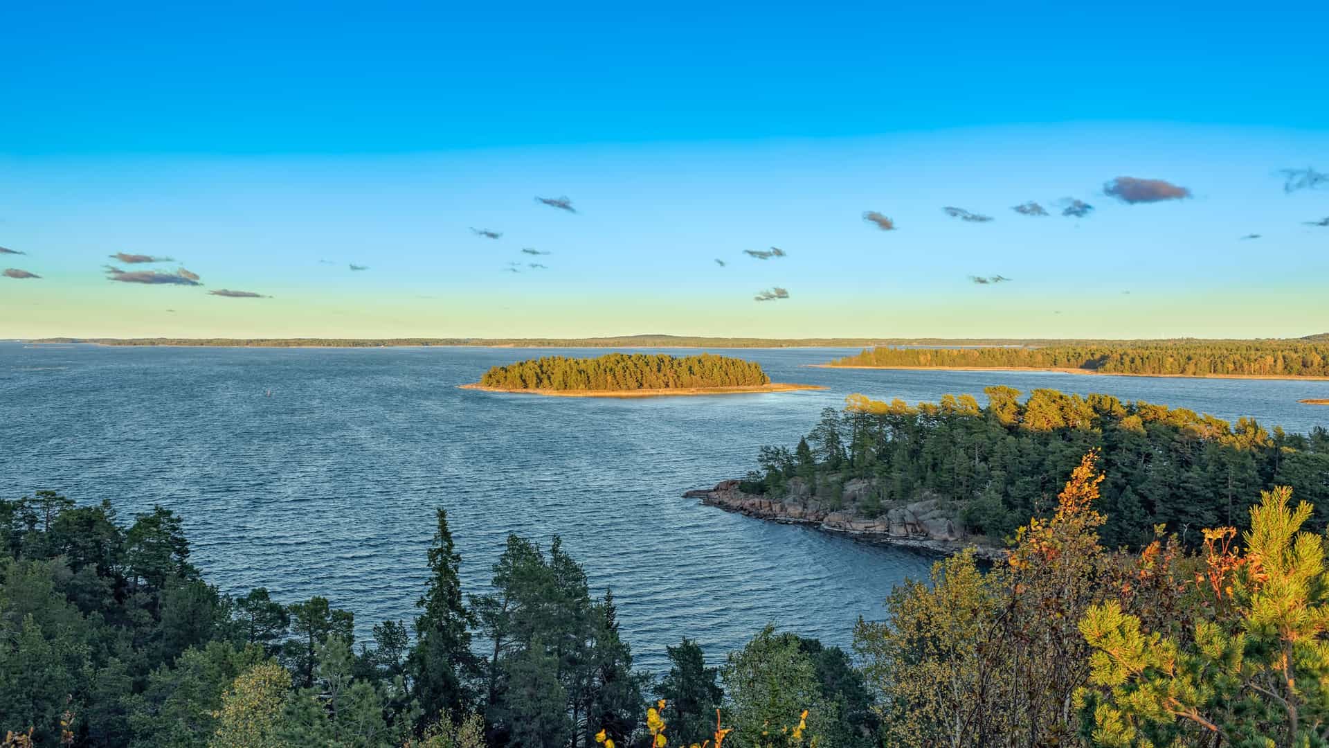 A beautiful evening landscape of the Gulf of Bothnia, with a wide expanse of water dotted with small, forested islands and a bright blue sky.