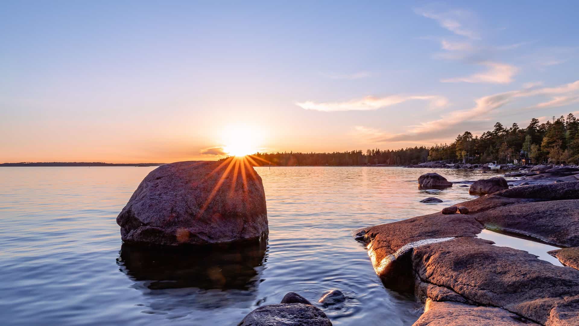 A beautiful sunset over the Gulf of Finland, with the sun's rays reflecting on the tranquil water and illuminating large boulders along a rocky shoreline.
