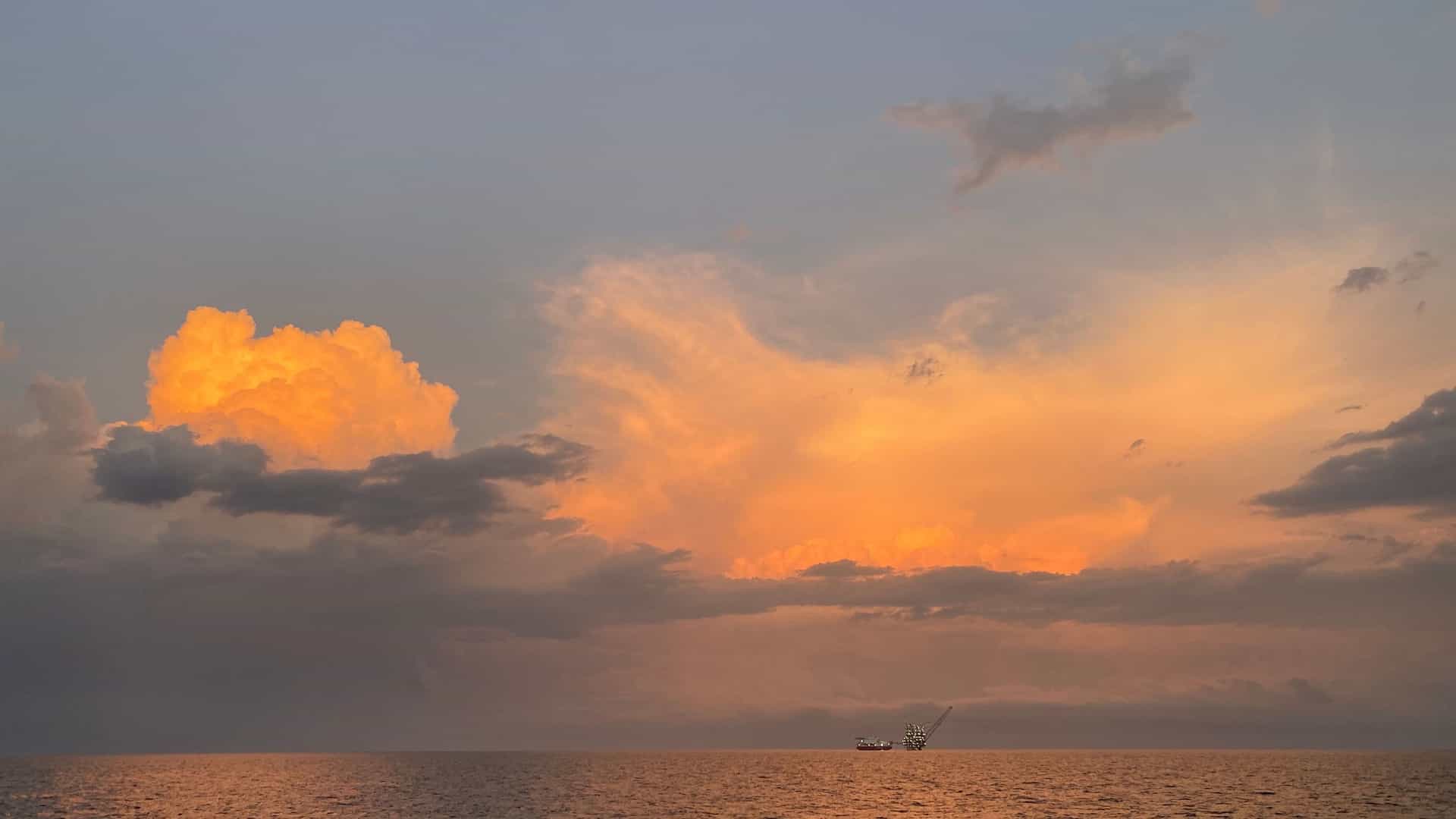 A beautiful, dramatic sunset over the Gulf of Guinea, with the sun's warm light illuminating large clouds and a distant oil platform on the calm sea.