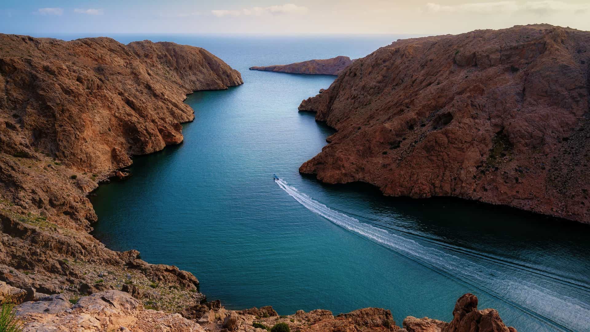 A scenic aerial view of a motorboat cruising through a narrow inlet of the Gulf of Oman, with high, dramatic rocky cliffs on both sides.