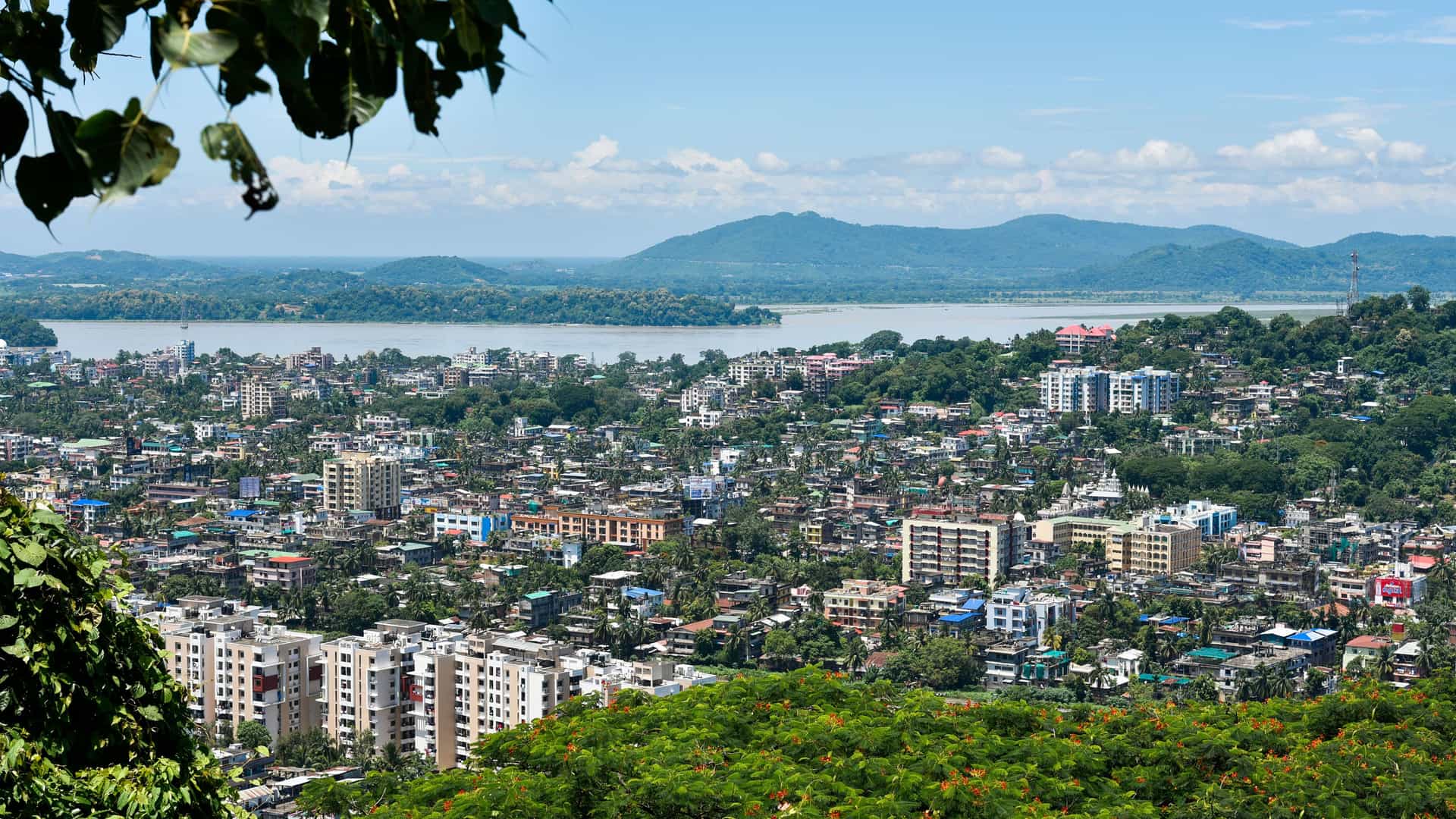"A scenic, elevated view of Guwahati, India, with the city's buildings nestled among lush trees, the Brahmaputra River in the background, and distant hills under a blue sky. "