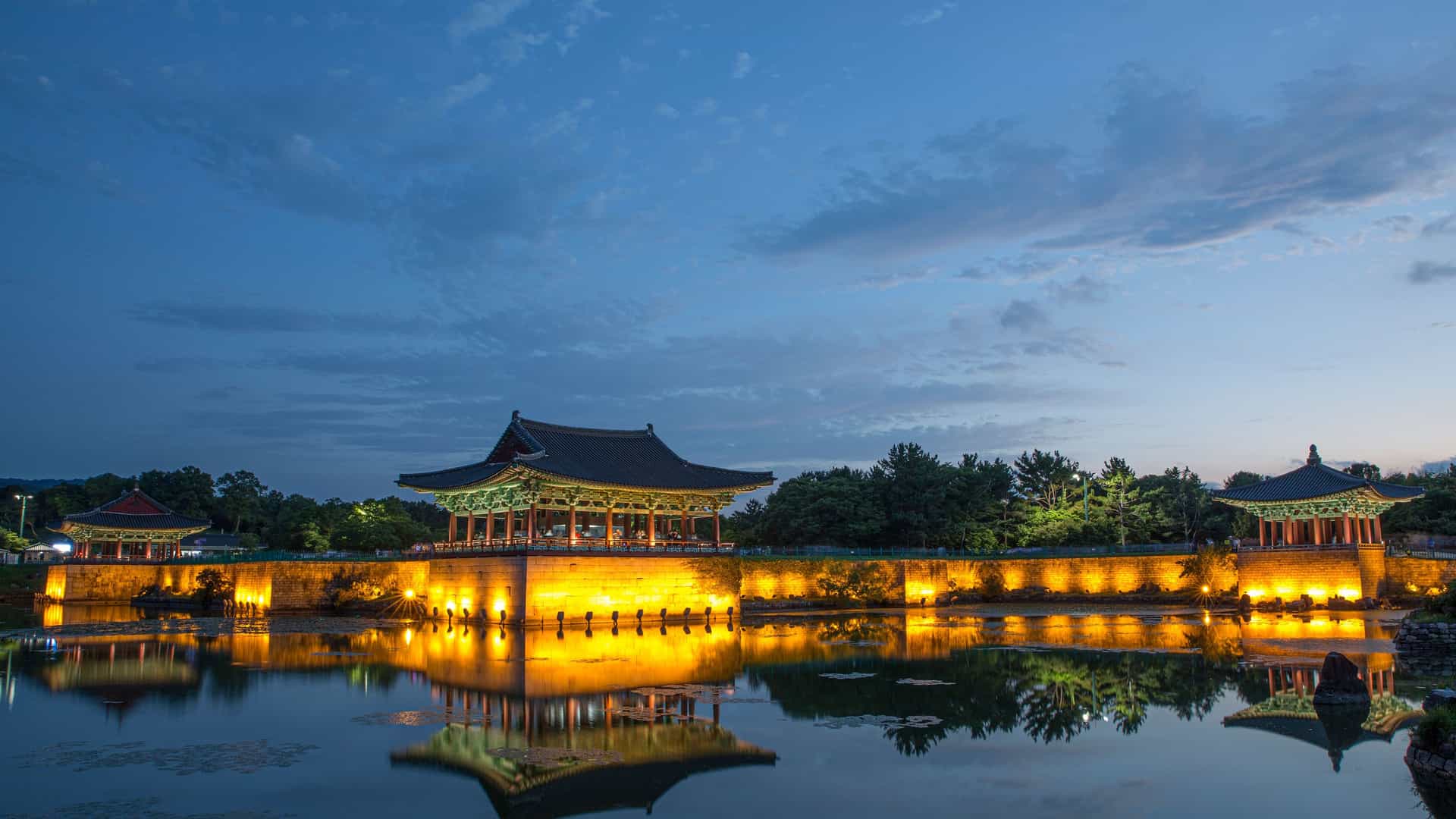 "A breathtaking night view of the Anapji Pond and Donggung Palace in Gyeongju, South Korea, with the brightly illuminated traditional buildings and their detailed reflections mirrored on the still water under a beautiful deep blue twilight sky. "