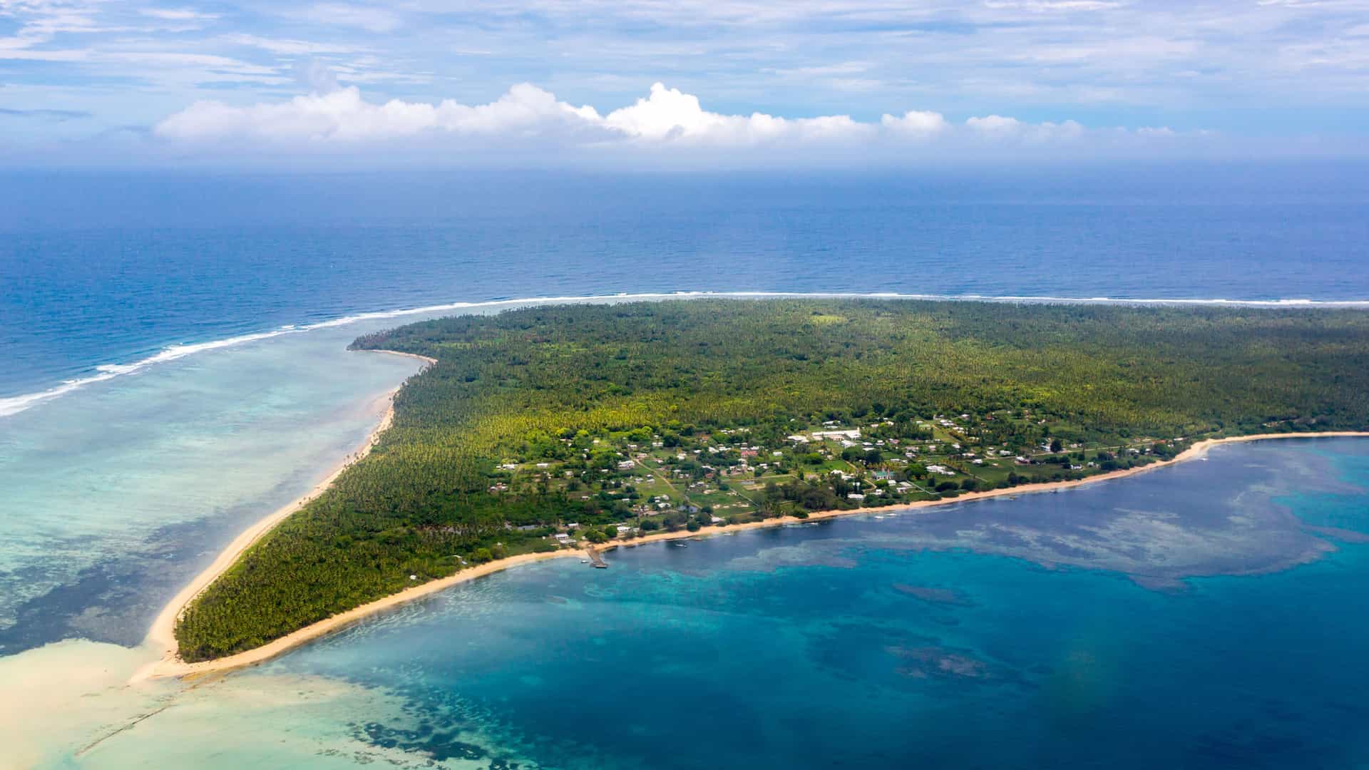 An aerial view of an island in the Ha'apai Group, Tonga, with a lush green interior and a village nestled between a white sand beach and turquoise water.