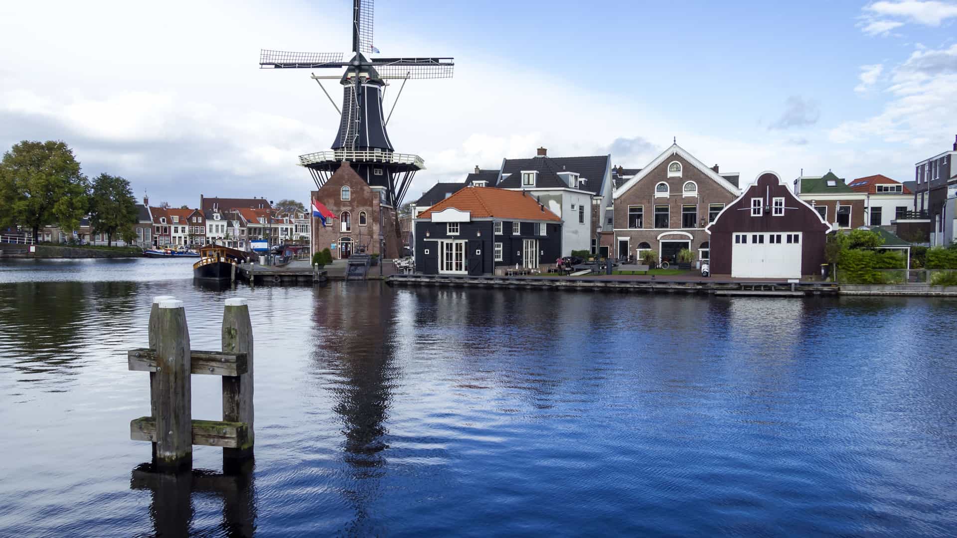 A picturesque view of Haarlem, Netherlands, with the De Adriaan windmill standing tall behind historic houses lining the tranquil Spaarne river under a cloudy sky.