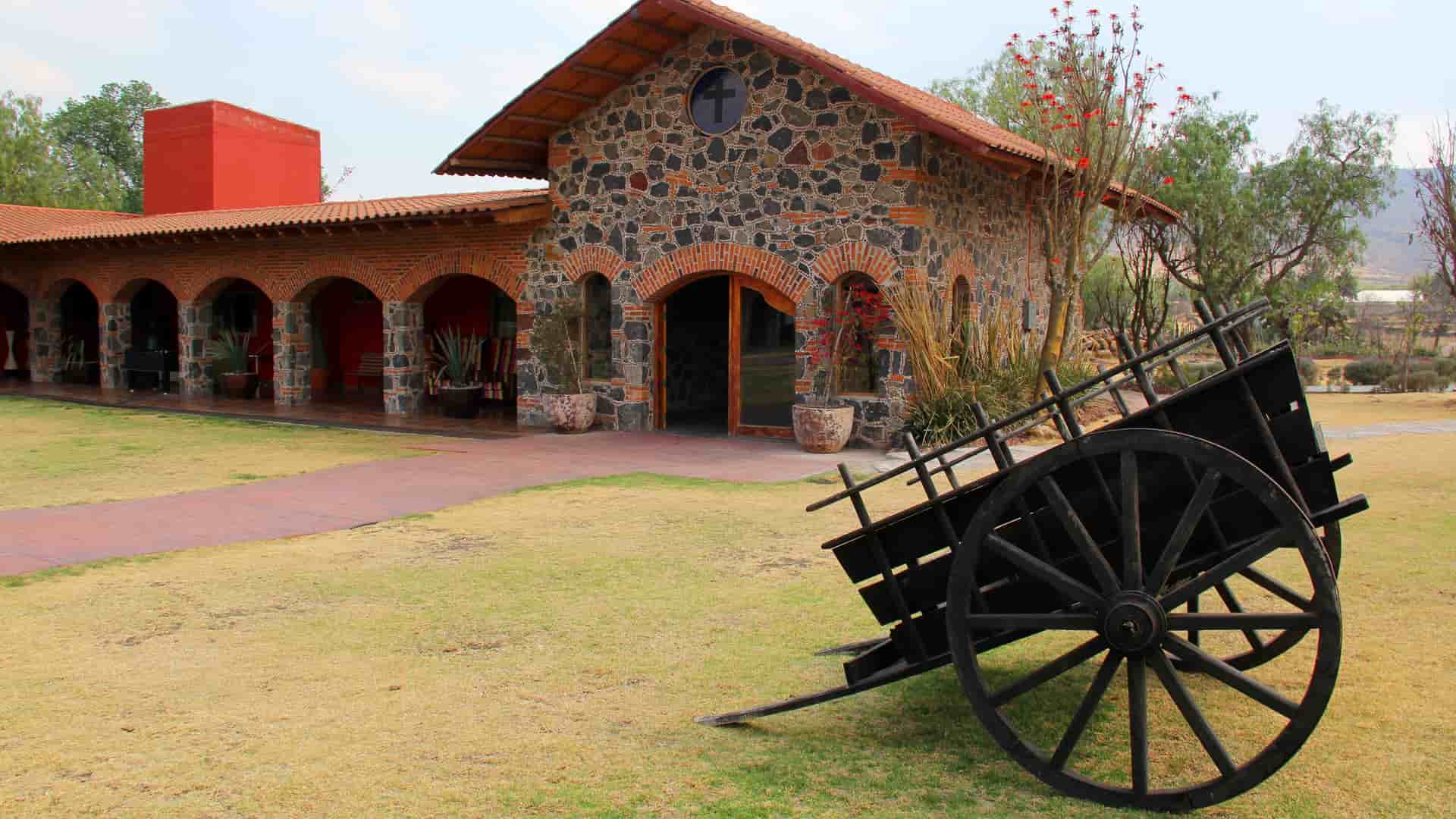 "A historic colonial-era building with a stone facade and red-tiled roof, part of the Hacienda Concepción in Peru, with a large, old wooden cart on the grassy lawn in front. "