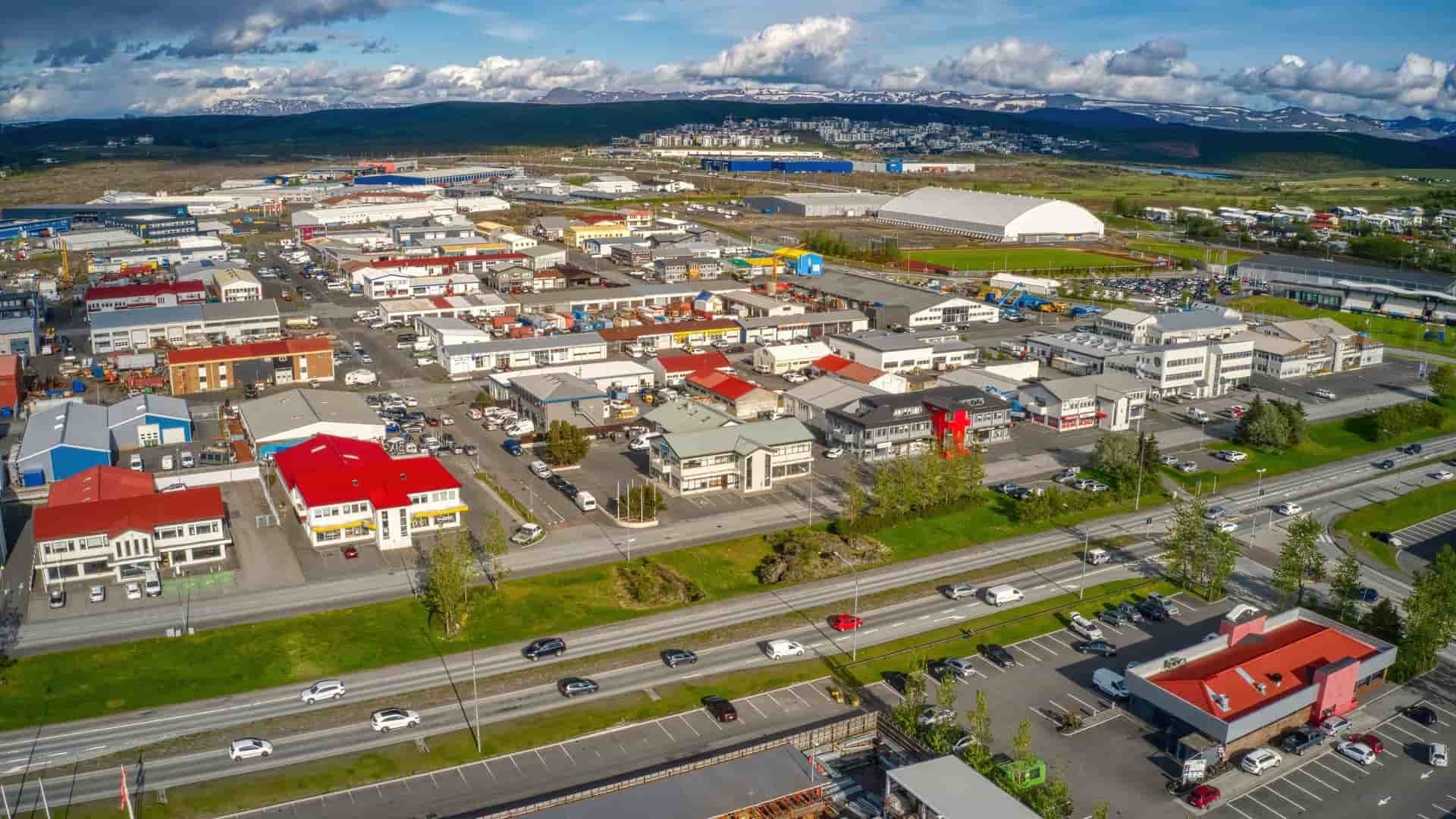 Aerial view of the industrial and commercial district of Hafnarfjörður, Iceland, showing a dense collection of low-rise buildings and warehouses, with a busy main road, against a backdrop of distant, snow-capped mountains.