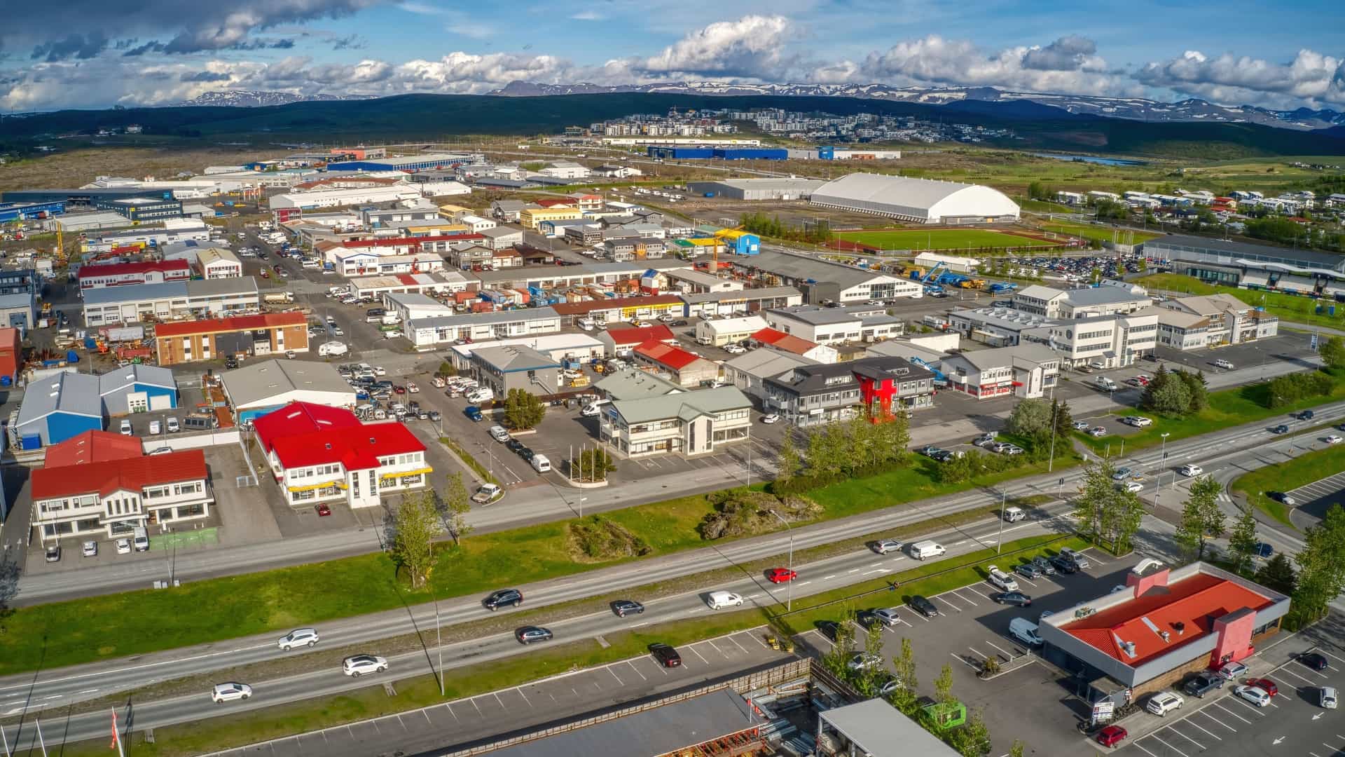 Aerial view of the industrial and commercial district of Hafnarfjörður, Iceland, showing a dense collection of low-rise buildings and warehouses, with a busy main road, against a backdrop of distant, snow-capped mountains.