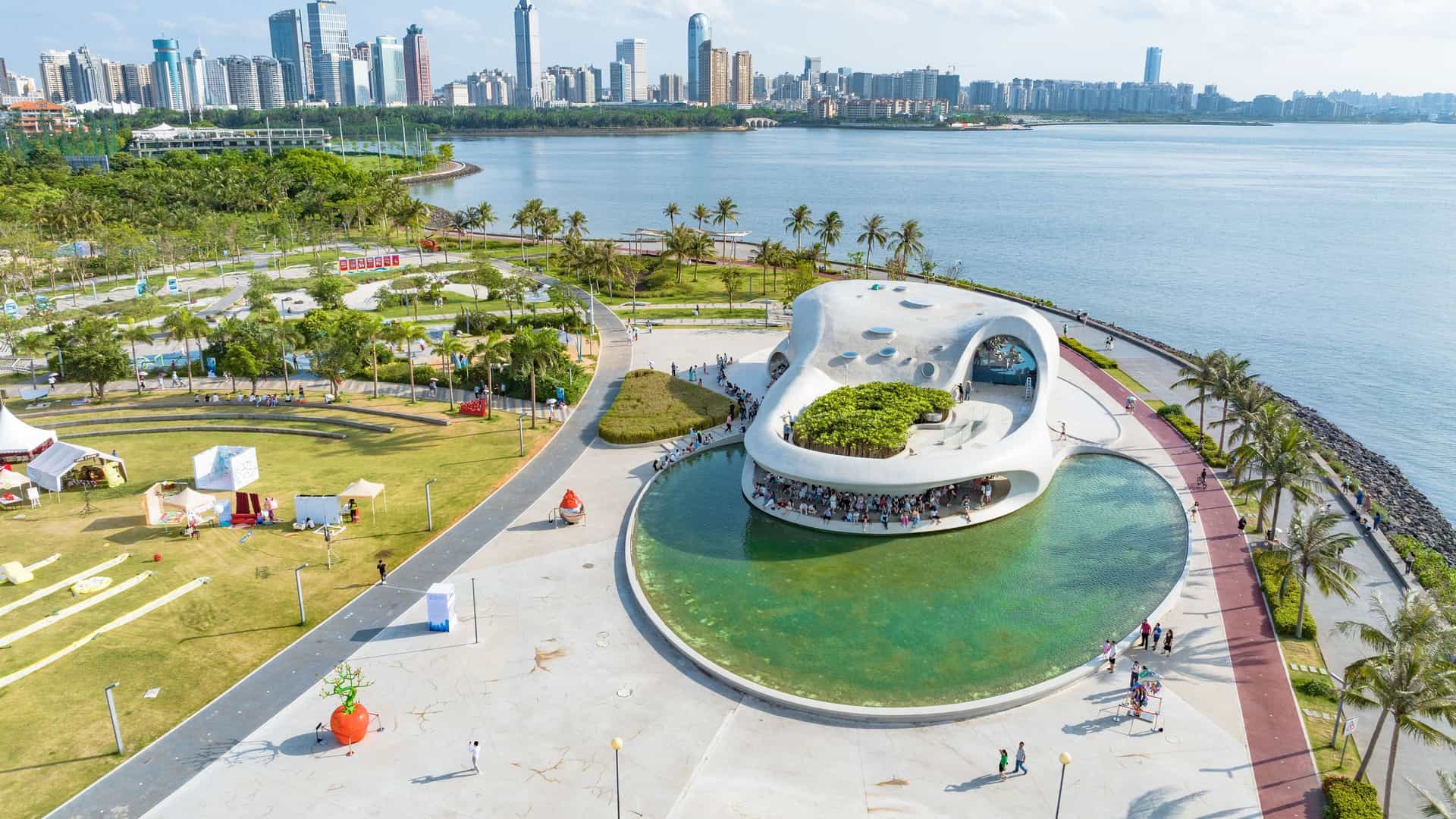 An aerial view of a modern building with a unique, shell-like design in Haikou, China, with a waterfront promenade, green parks, and a skyline in the distance.