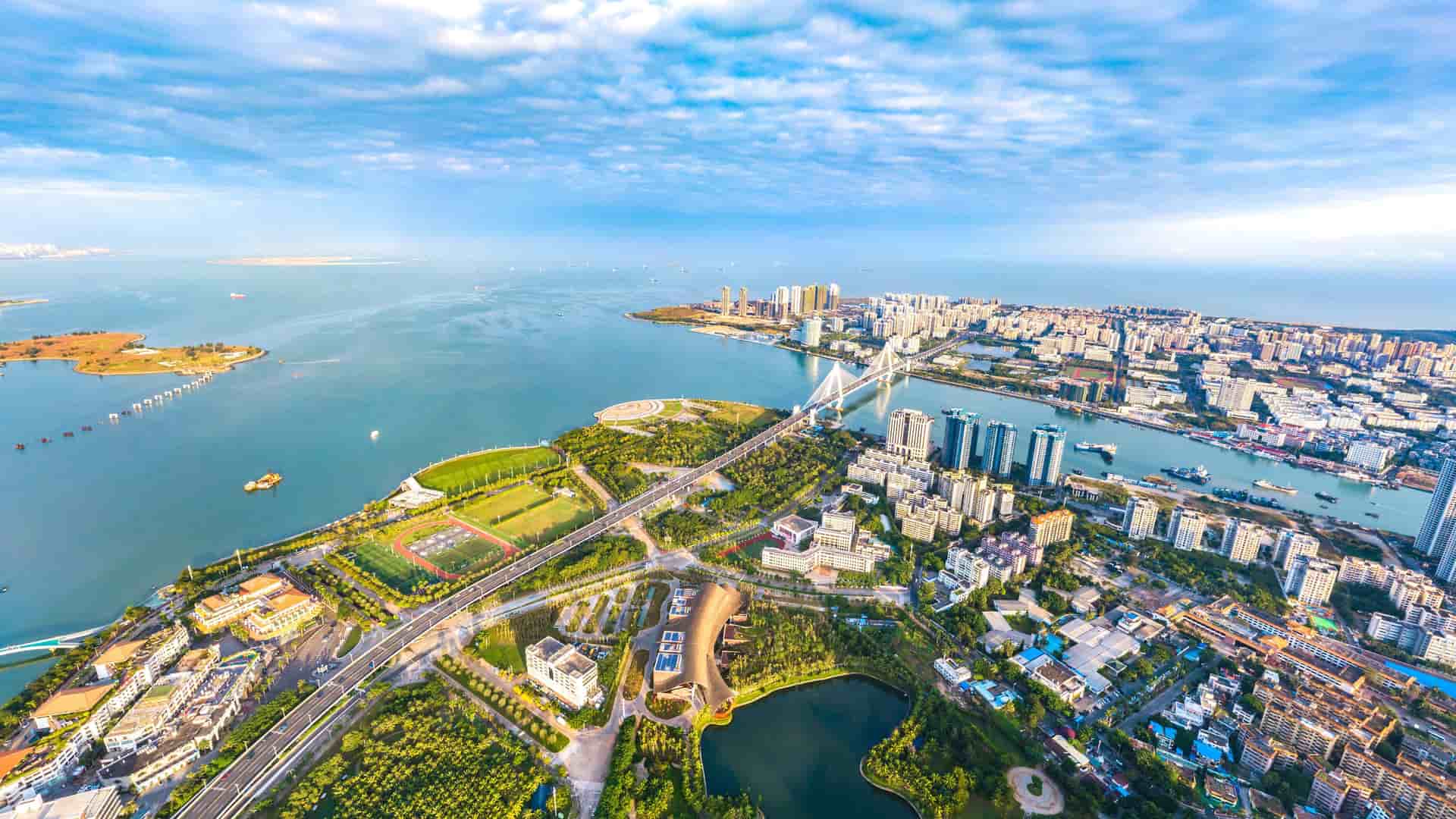 An aerial view of the Hainan Strait, showing a sprawling modern city with a large bridge connecting a green, coastal area to a dense urban center, all under a blue sky.
