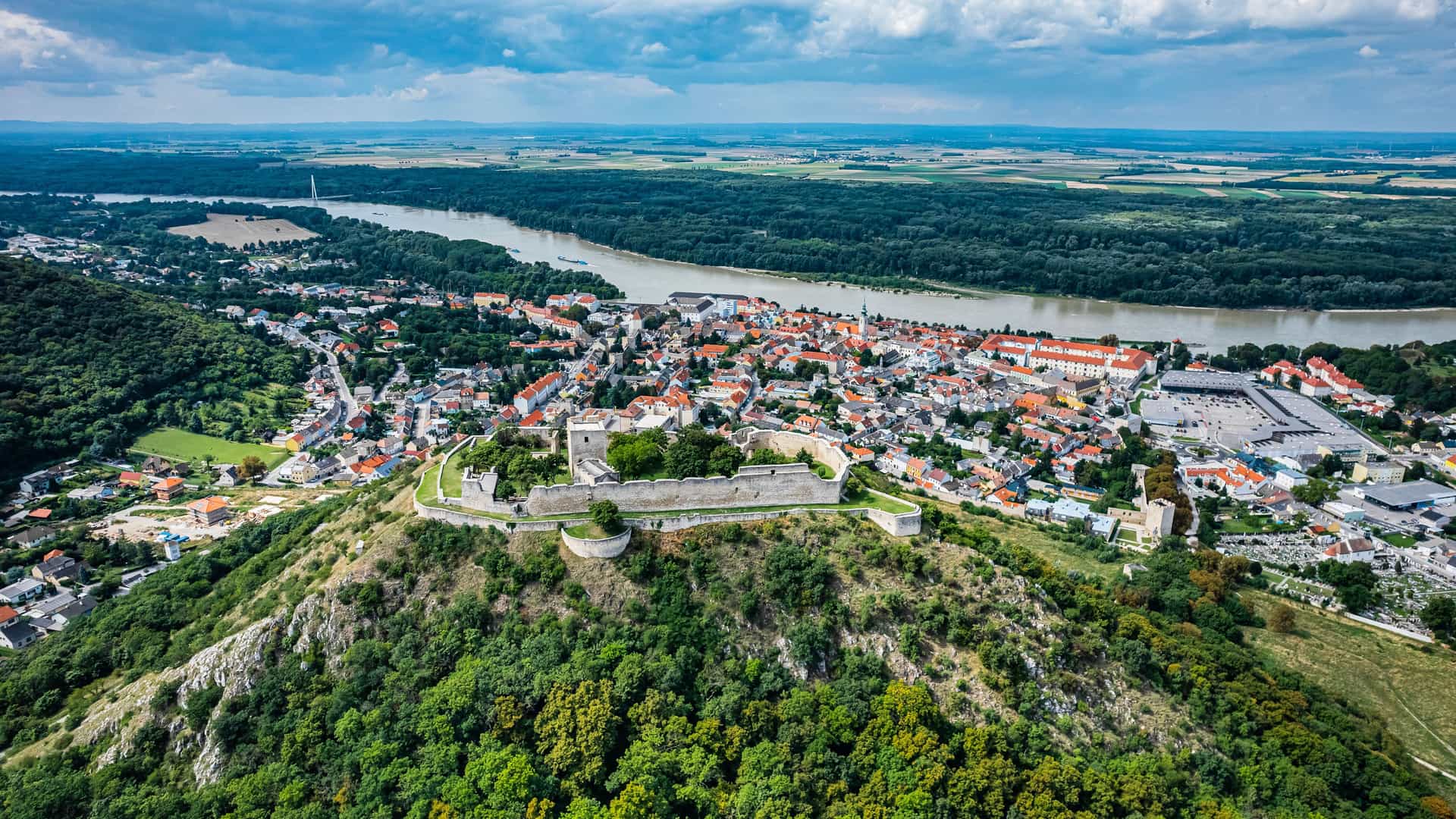 "An aerial view of Hainburg, Austria, with the ruins of the historic Hainburg Castle perched on a hill overlooking the town and the vast Danube River winding through the green landscape. "