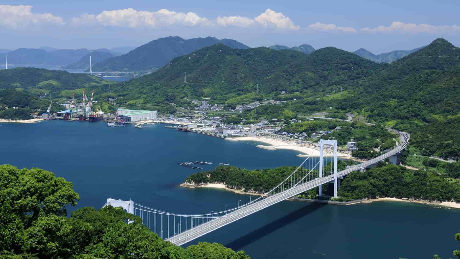 A high-angle view of a suspension bridge spanning a bay near Hakata, Japan, with green, forested mountains in the background and a town with a small harbor along the shore.