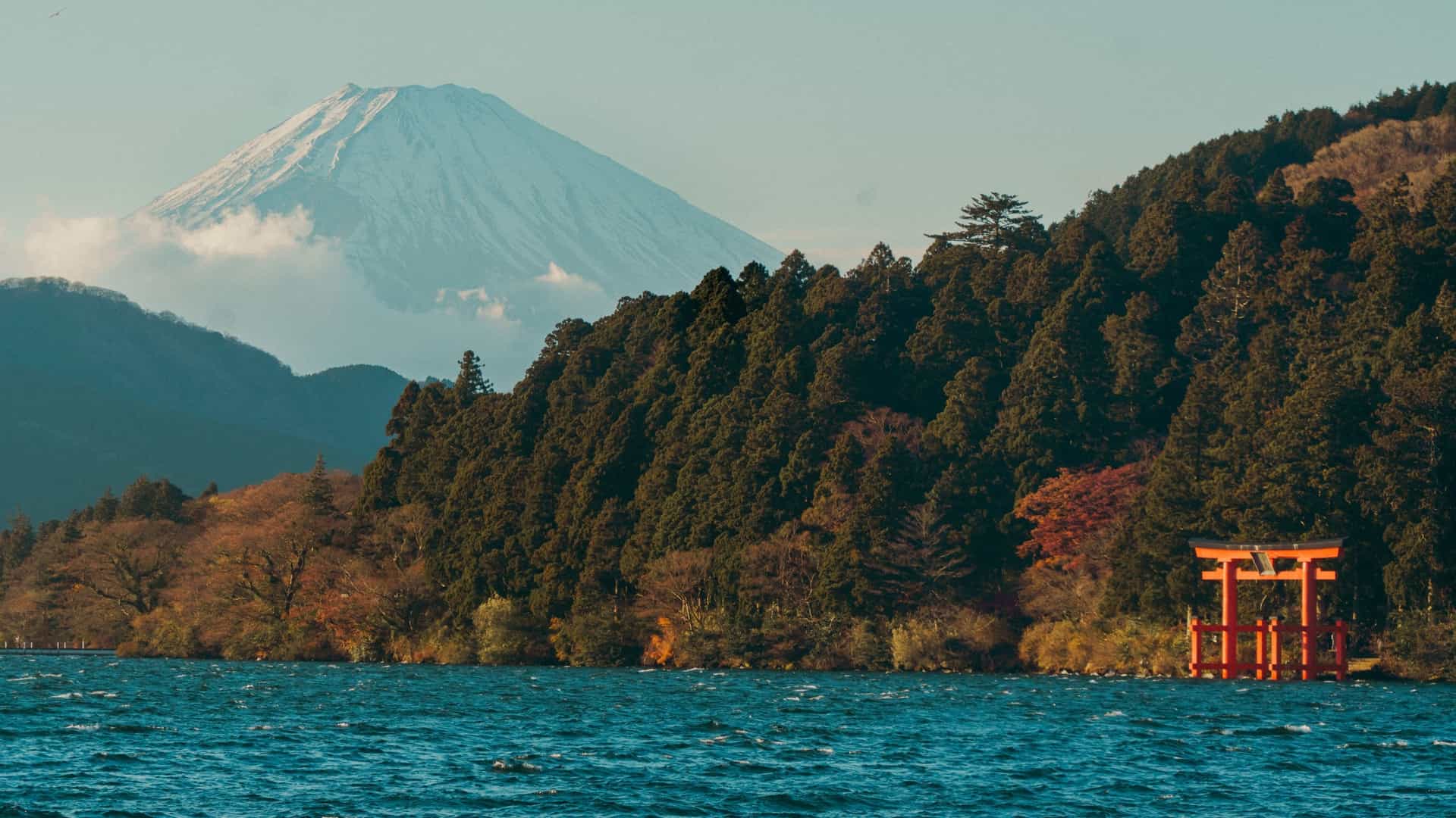 A scenic view across Lake Ashi in Hakone, Japan, with the iconic snow-capped peak of Mount Fuji visible in the background over a dense, pine-covered hillside, and a bright orange torii gate standing at the water's edge.