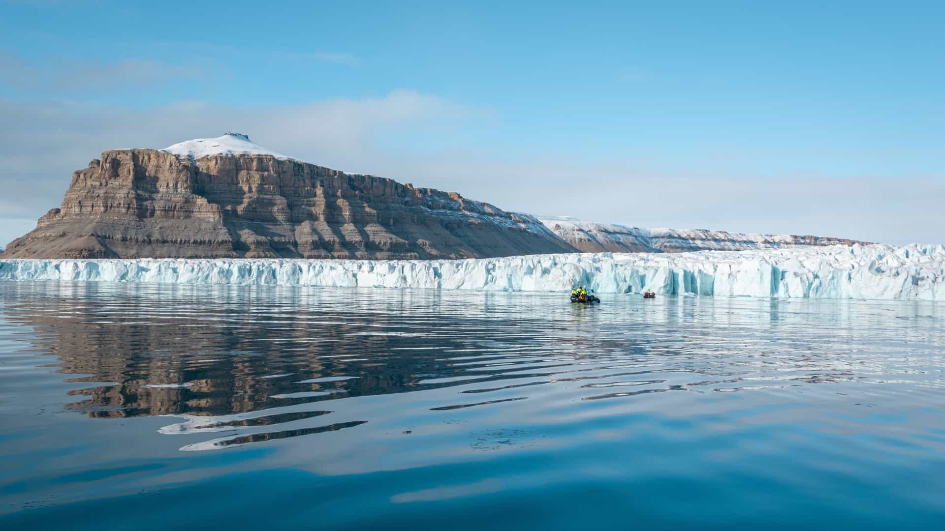 A dramatic polar scene in Halford Island with a towering glacier meeting calm icy water, a rugged snow‑covered mountain rising behind it, and a small boat of people in bright gear emphasizing the immense scale of the landscape.