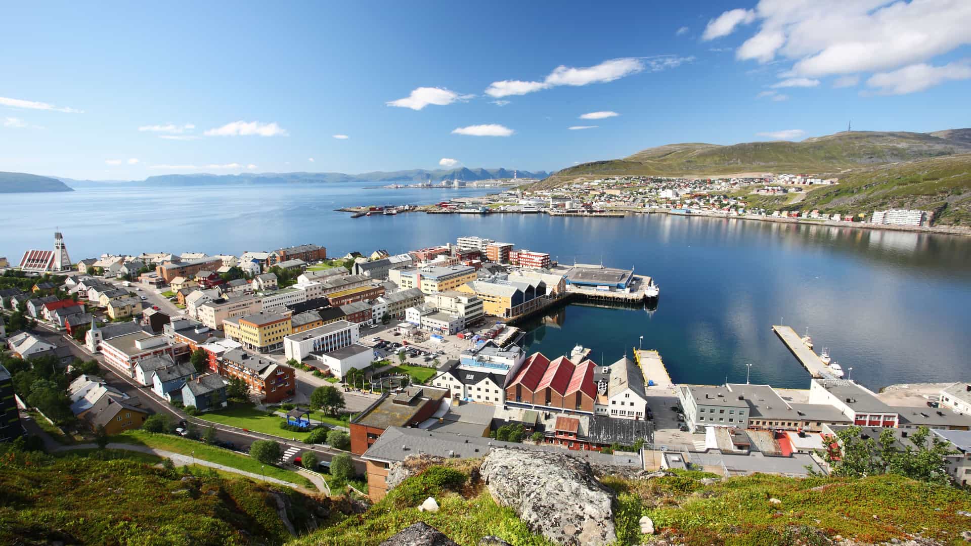 A scenic high-angle view of the harbor and town of Hammerfest, Norway, with colorful buildings and a fjord in the background under a blue sky with white clouds.