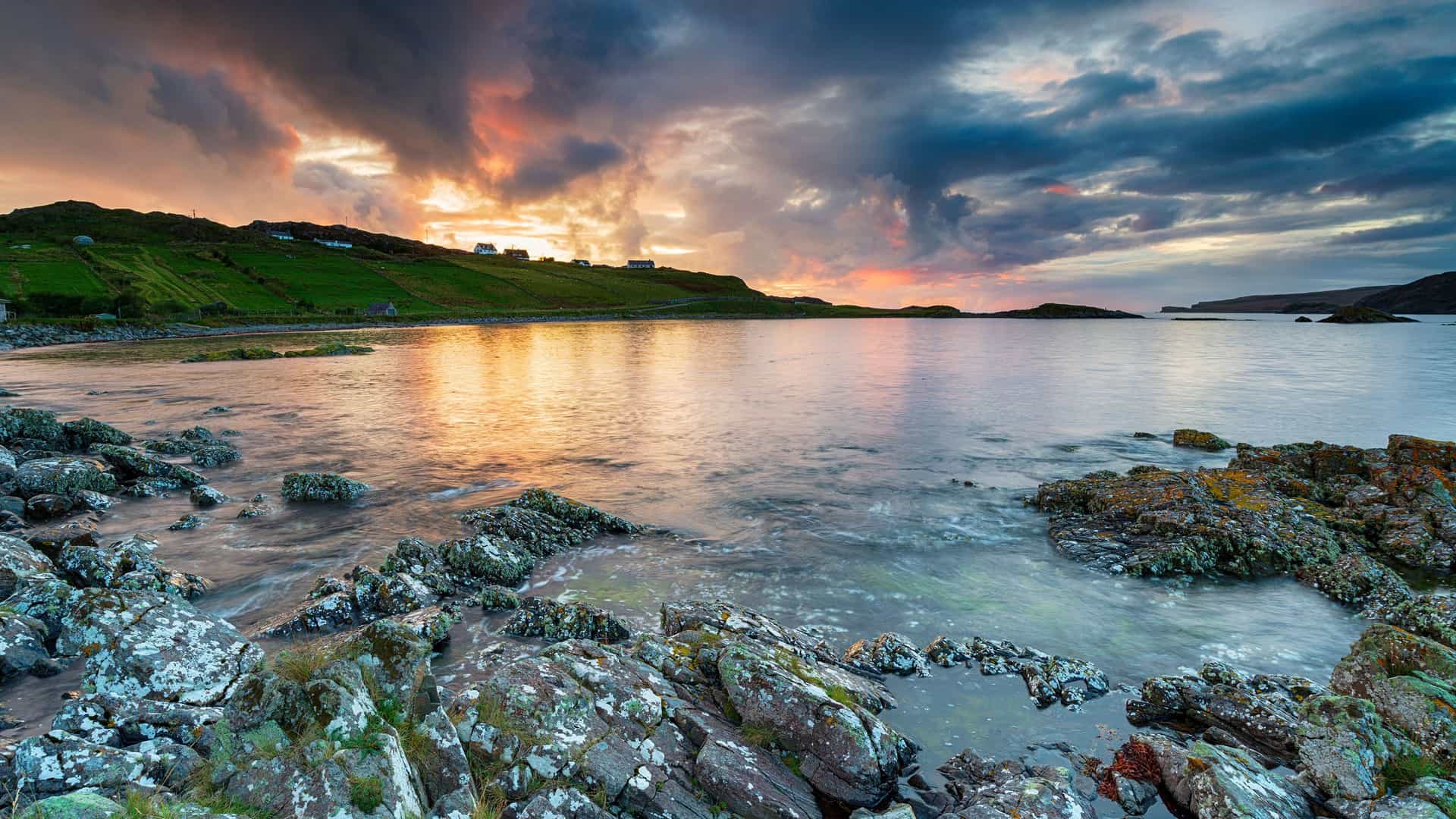A dramatic sunset over Handa Island, Scotland, with the sky ablaze in orange and pink hues, reflecting on the tranquil water and illuminating the rocky shore.