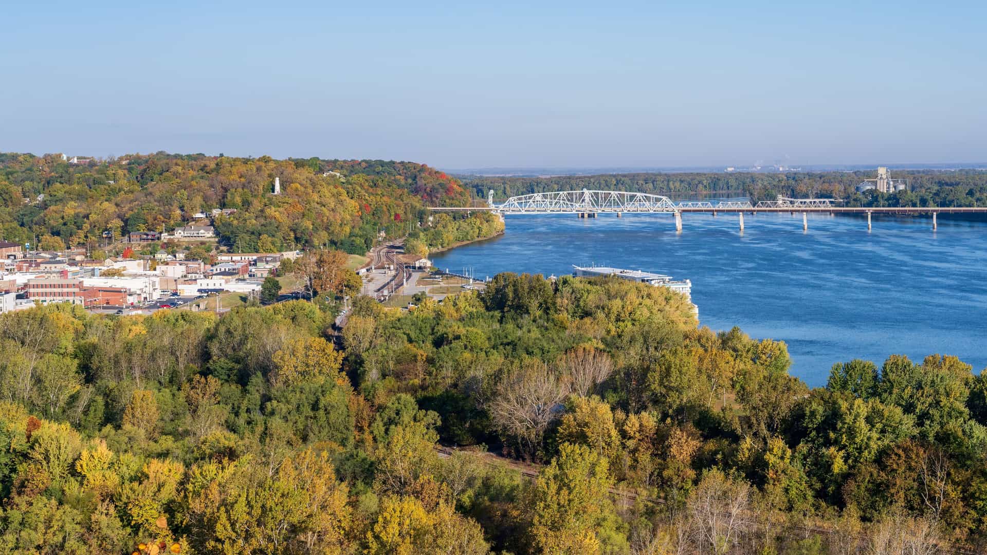A panoramic, elevated view of Hannibal, Missouri, with the city nestled along the Mississippi River, with a bridge spanning the water and colorful trees in the foreground.