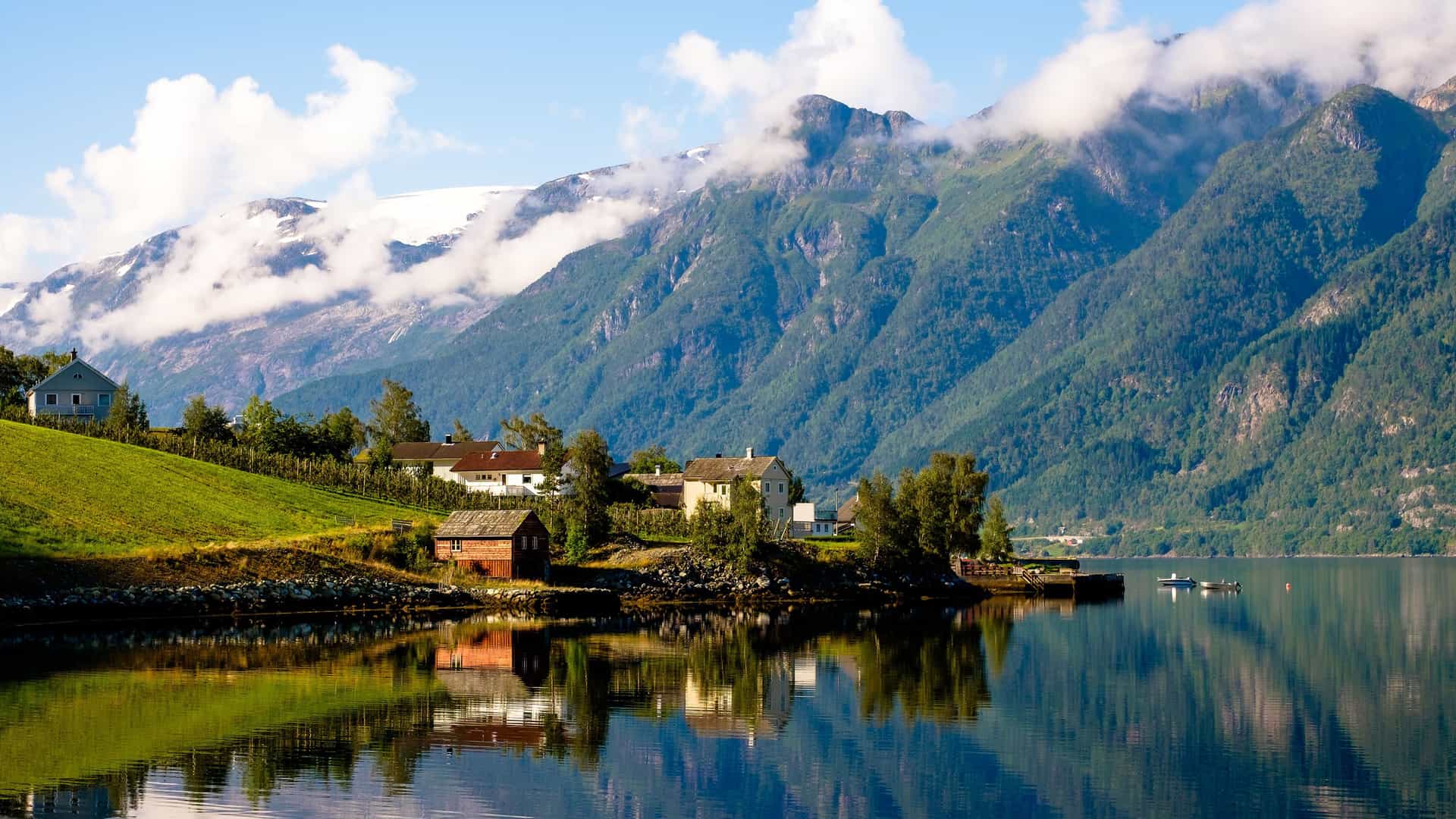 A picturesque view of Hardangerfjord, Norway, with small houses on a lush, green hillside reflected in the calm water and towering snow-capped mountains in the background.
