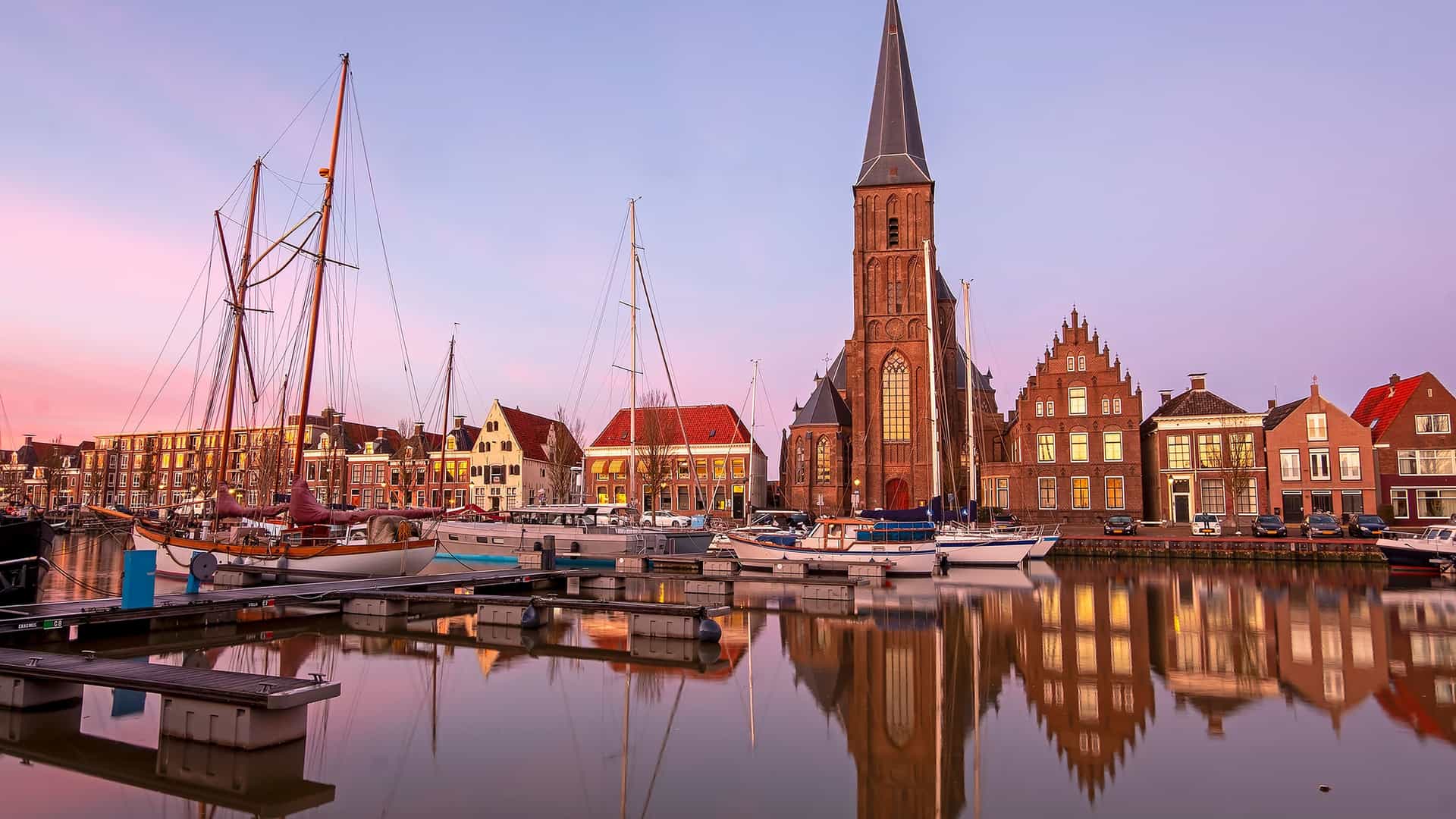 A picturesque sunset over Harlingen, Netherlands, with the majestic Grote Kerk church and traditional houses reflected in the calm waters of the harbor filled with sailboats.