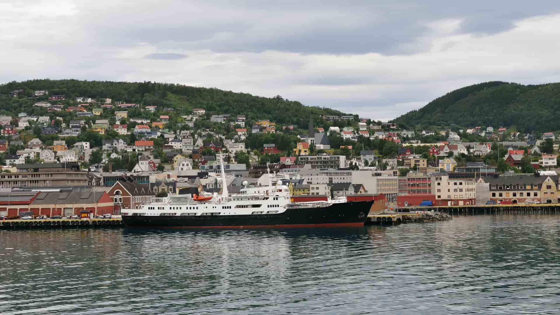 A large black and white ship is docked at the harbor in Harstad, Norway, with the city's colorful houses sprawling up the lush green hillsides in the background.