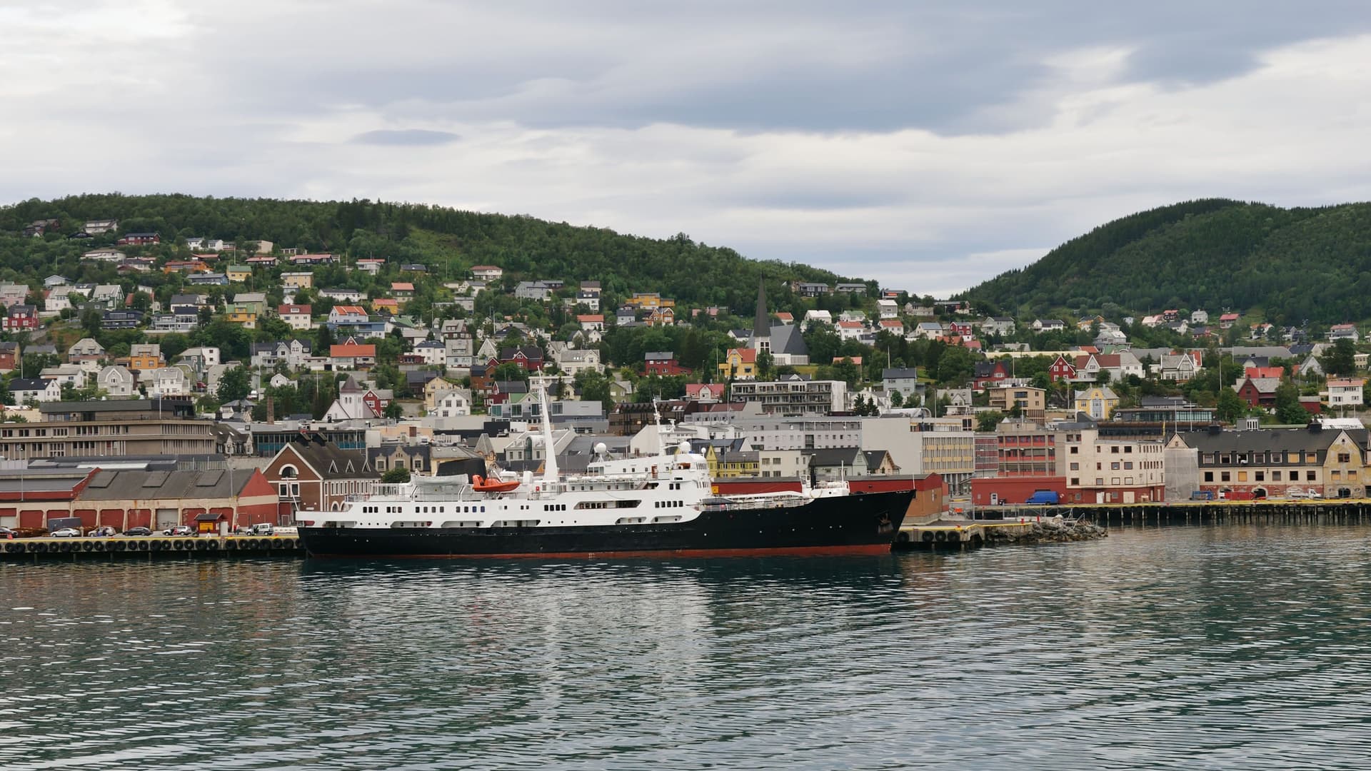 A large black and white ship is docked at the harbor in Harstad, Norway, with the city's colorful houses sprawling up the lush green hillsides in the background.