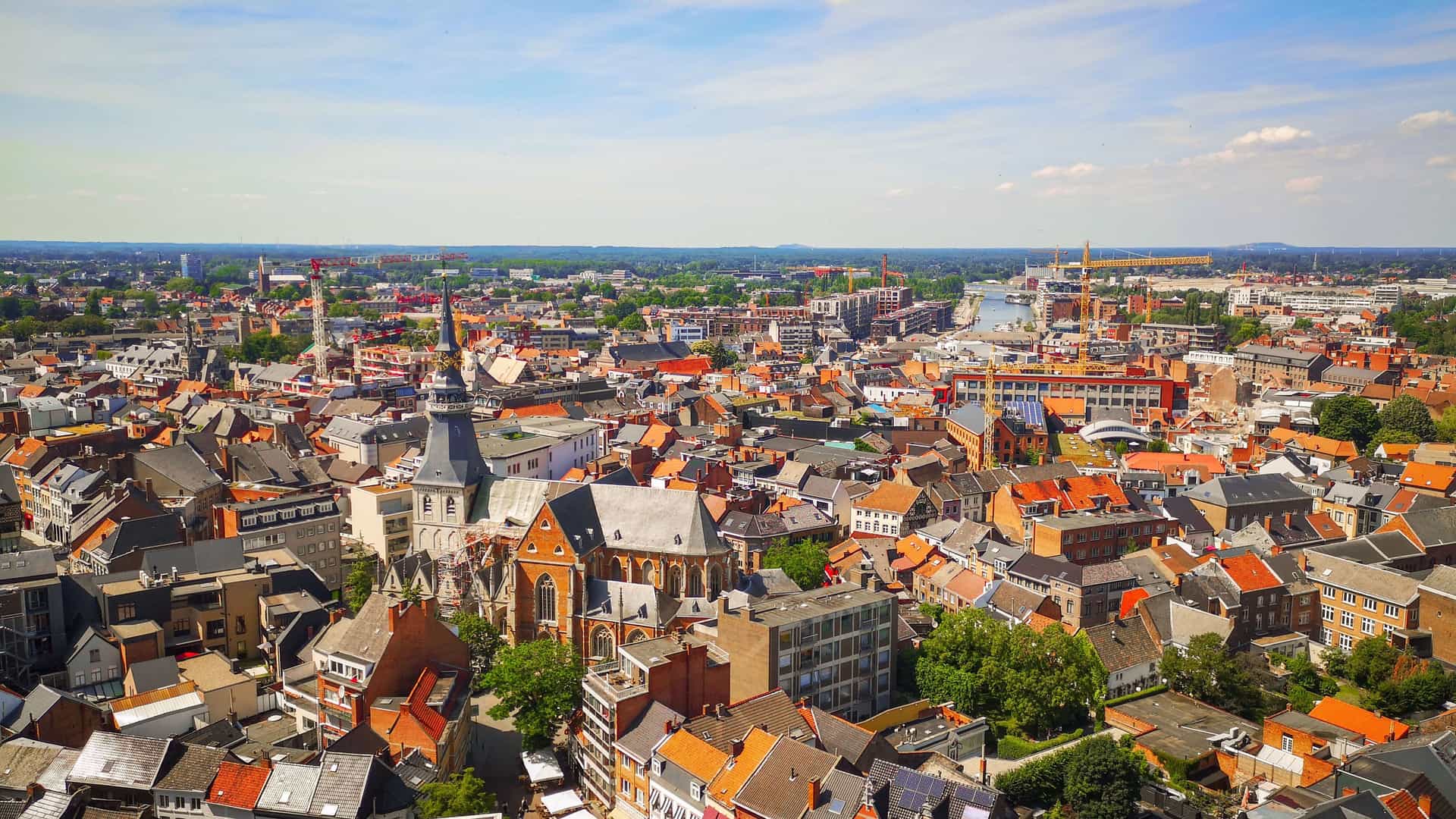 An aerial view of Hasselt, Belgium, showing a densely packed cityscape with many buildings featuring traditional orange-tiled roofs and a river winding through the urban landscape.
