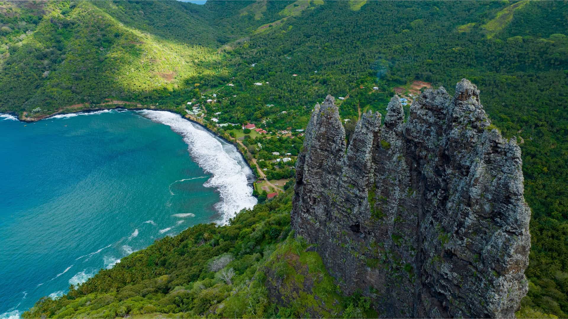 An aerial view of Hatiheu Bay in the Marquesas Islands, with a stunning black sand beach, a small village, and a large, jagged rock formation towering over the bay.