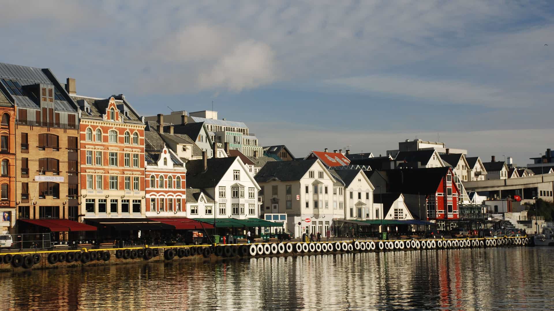 A view of the waterfront in Haugesund, Norway, with traditional wooden houses and cafes lining the harbor, reflected in the still water on a partly cloudy day.