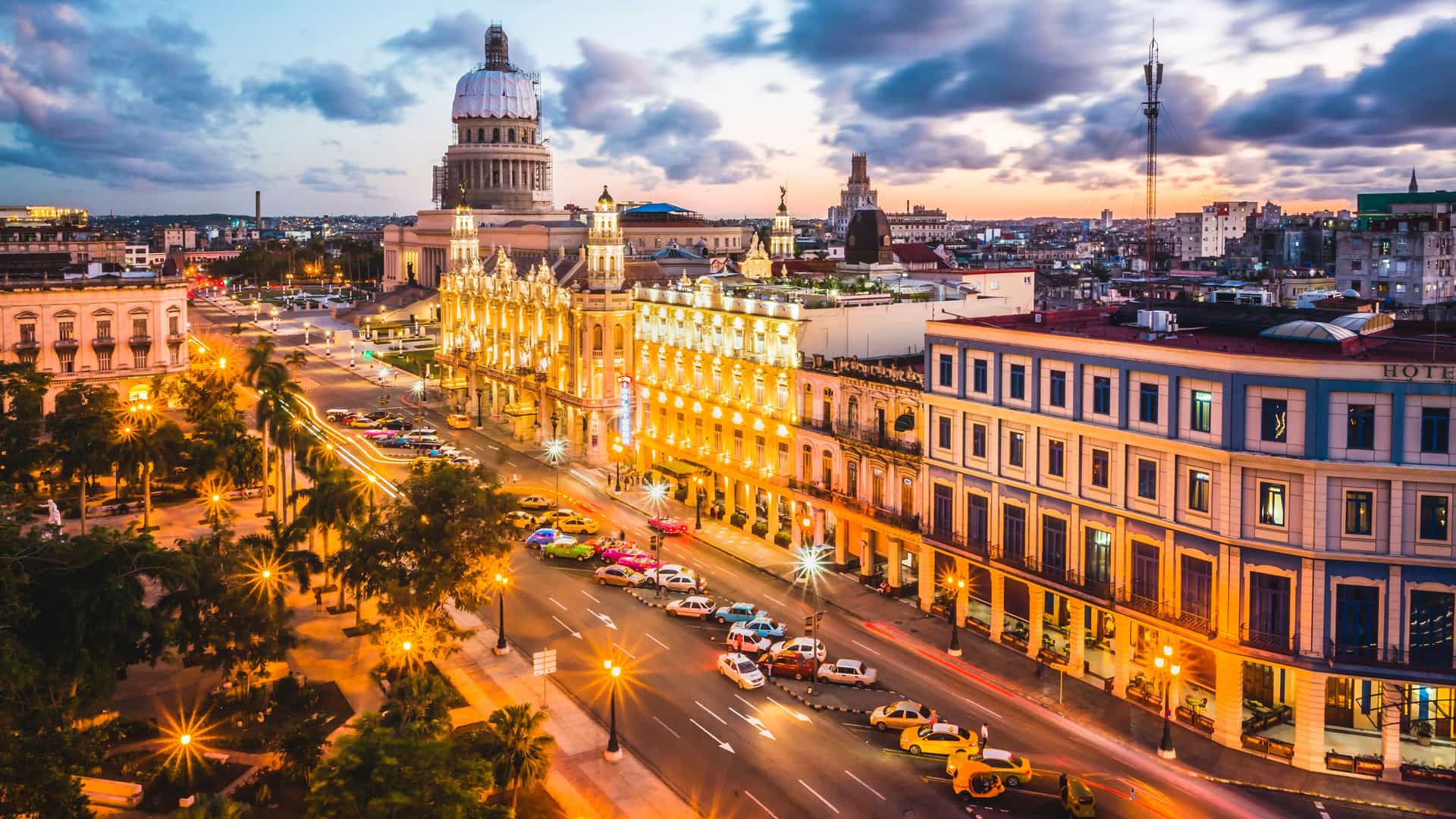 "A wide-angle, elevated view of downtown Havana, Cuba at sunset, showing the iconic Capitolio building, classic cars on the bustling street, and glowing streetlights. "