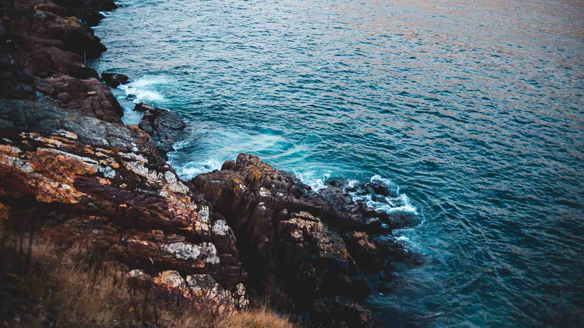 A rugged, high-angle shot of the dramatic rocky coastline of Hebron, Labrador, with waves crashing against the jagged cliffs and dark blue water stretching to the horizon.