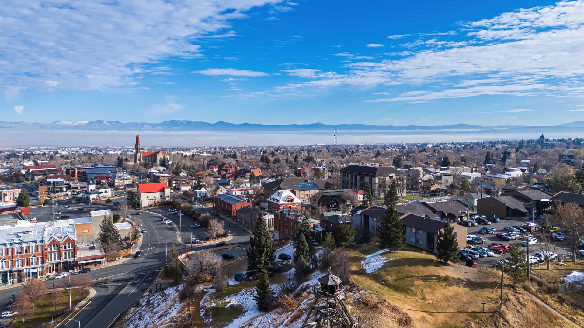 "An aerial view of Helena, Montana, with the city's historic buildings and the iconic Cathedral of St. Helena in the foreground, and the snow-capped mountains of the Rocky Mountains in the distance under a blue sky. "
