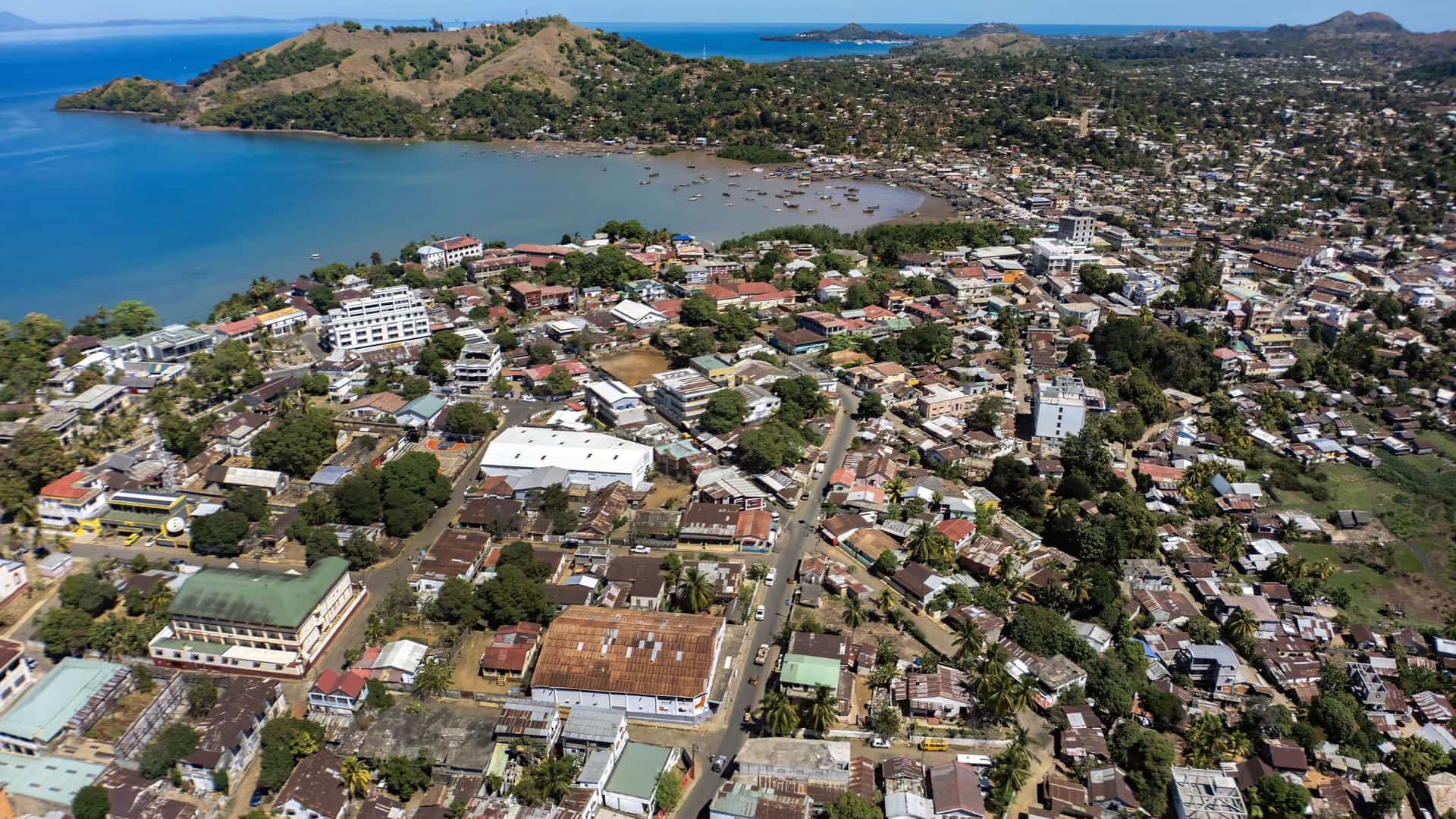 An aerial view of Hell-Ville, Madagascar, showing a dense, sprawling town along a beautiful crescent-shaped bay, with a harbor filled with small boats and lush, green hills in the background.