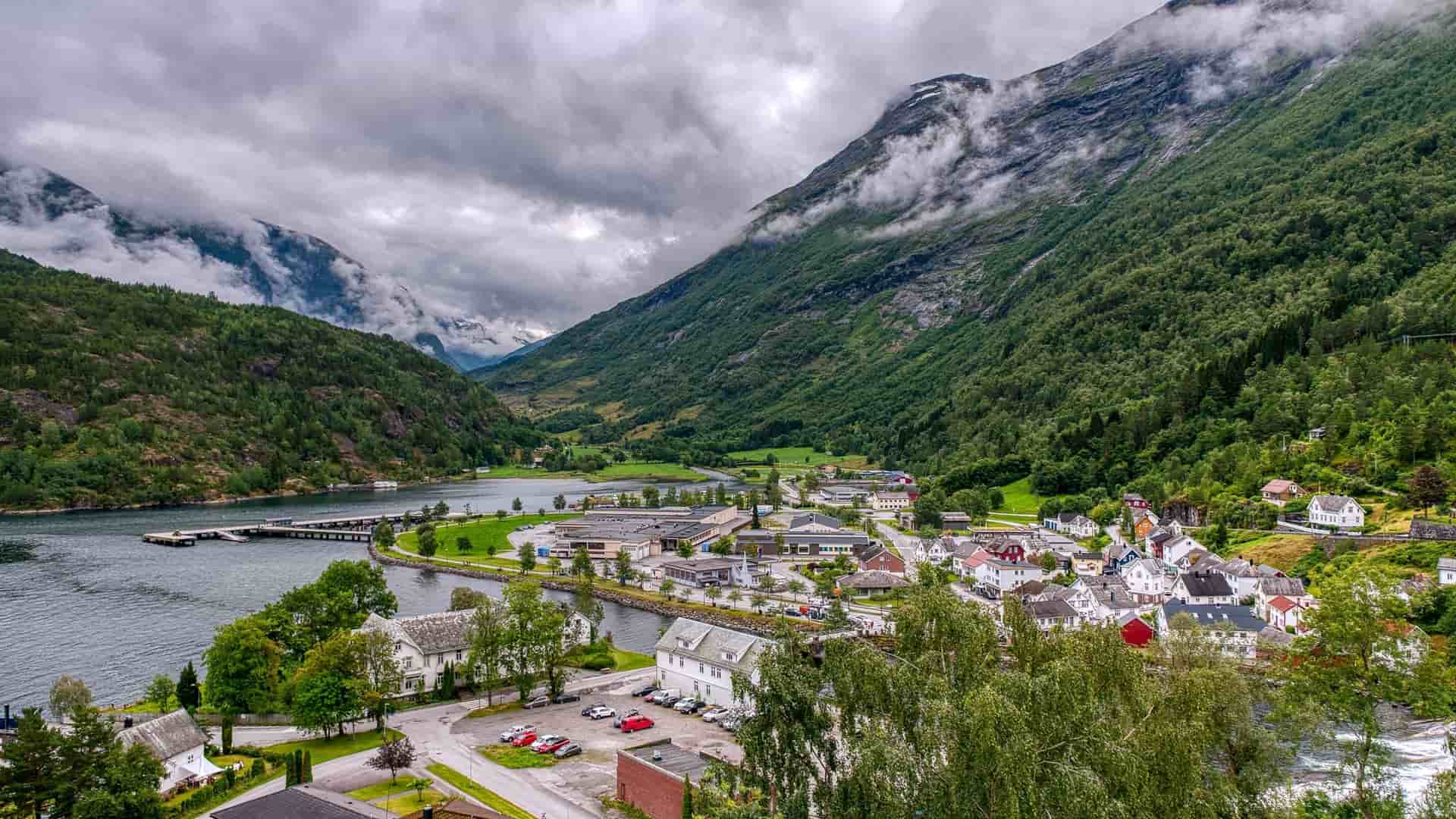 An aerial or high-angle view of the scenic Hellesylt village in Stranda municipality, Norway, situated at the head of the Sunnylvsfjord. The village is characterized by traditional Norwegian buildings, surrounded by steep, lush green mountains, with the powerful Hellesylt Waterfall prominently dividing the settlement as it flows into the fjord.