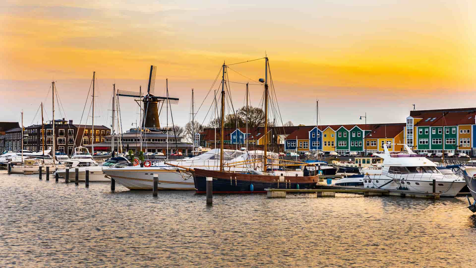 A stunning sunset over Hellevoetsluis, Netherlands, with a bustling harbor filled with sailboats and yachts, and a traditional windmill and colorful houses lining the waterfront.