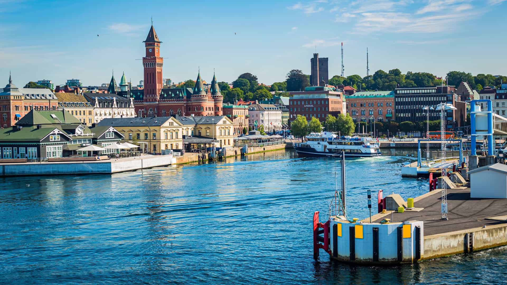 "A sunny panoramic shot of the Helsingborg waterfront in Sweden, with the city's charming architecture and historic clock tower reflected in the harbor's calm, blue water as a ferry approaches the dock. "