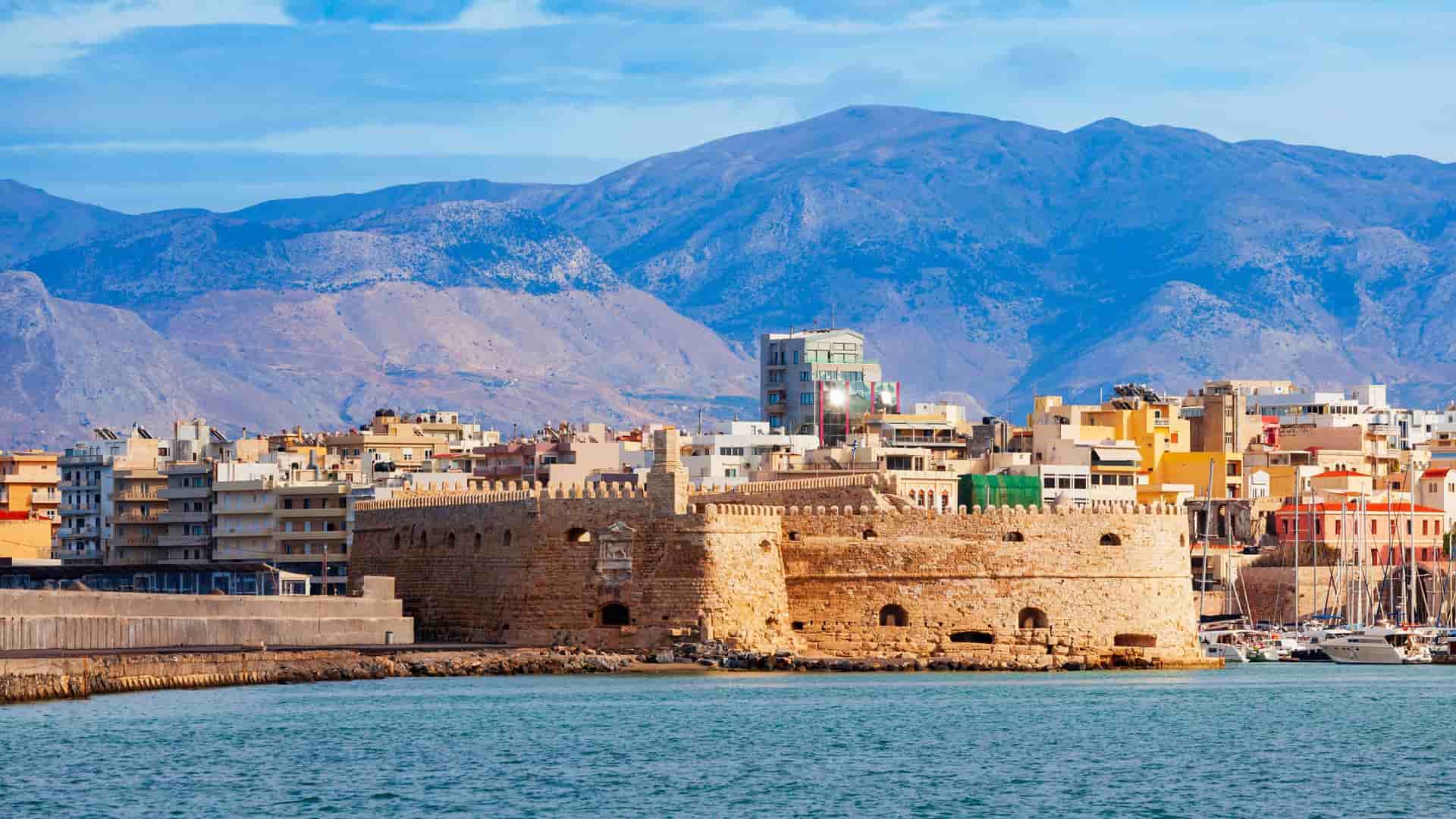 A picturesque view of Heraklion, Crete, with the historic Koules Fortress in the foreground, the bustling port, and the city sprawling at the foot of a large, arid mountain range in the background.
