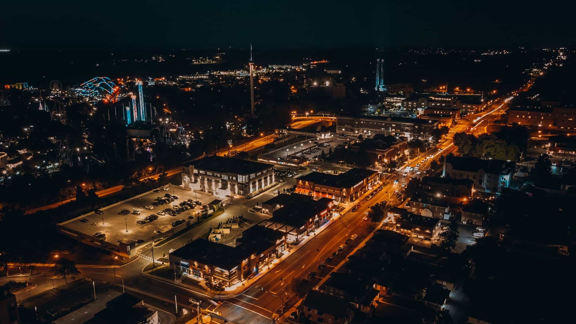 A vibrant nighttime aerial view in Hershey featuring brightly lit streets, a busy parking area, and a colorful amusement park with roller coasters and a Ferris wheel glowing against the surrounding cityscape.