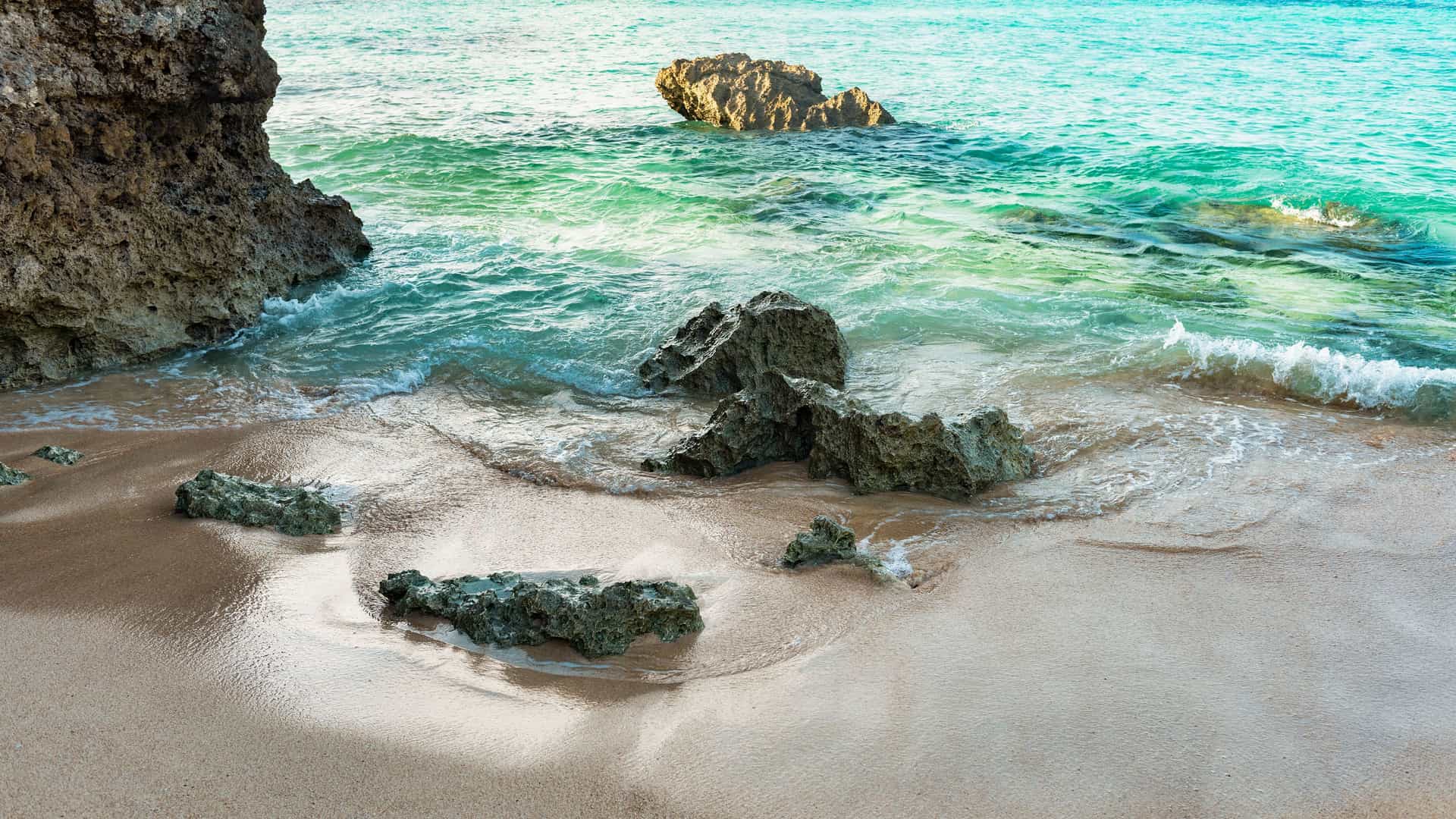 A tranquil view of a sandy beach in Hirara, Miyakojima, with gentle waves from the turquoise water lapping at the shore, and several large rocks scattered on the sand and in the water.