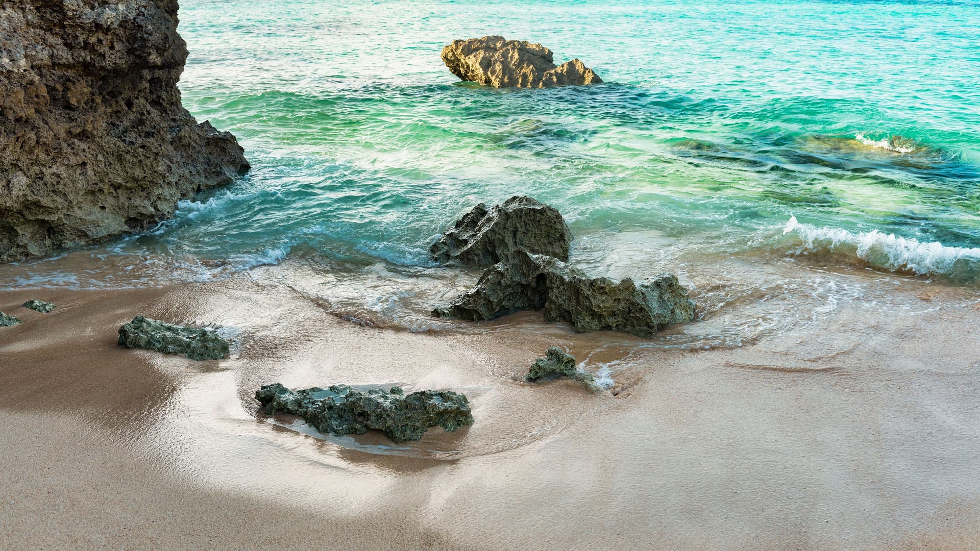 A tranquil view of a sandy beach in Hirara, Miyakojima, with gentle waves from the turquoise water lapping at the shore, and several large rocks scattered on the sand and in the water.