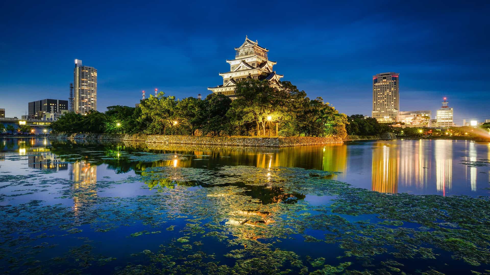 A stunning nighttime view of Hiroshima Castle in Japan, brightly lit and surrounded by a moat reflecting the castle and the modern city skyline in the background.