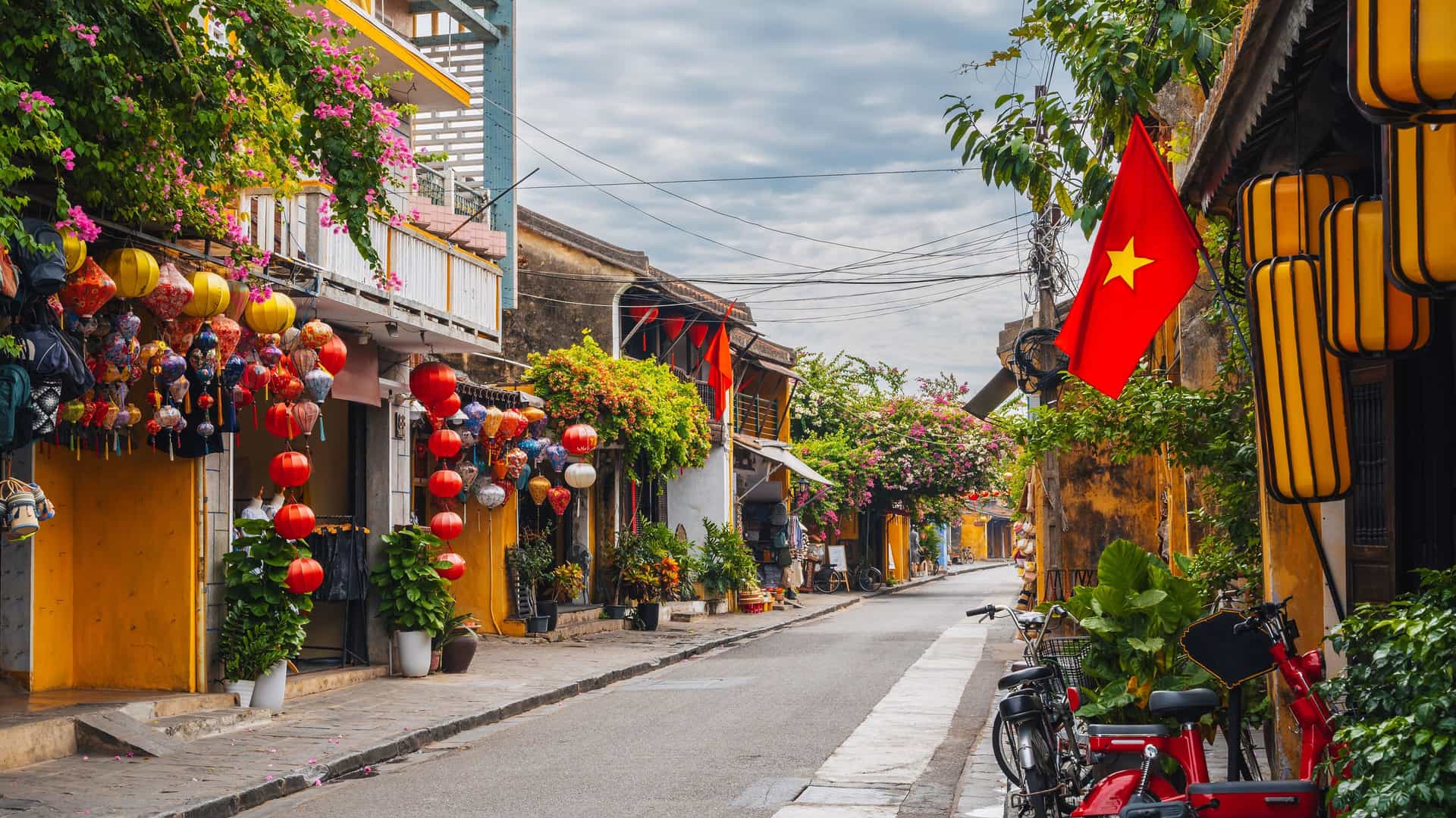 "A beautiful street in Hoi An, Vietnam, is lined with traditional yellow buildings adorned with colorful silk lanterns and blooming bougainvillea, with the Vietnamese flag flying from a storefront. "