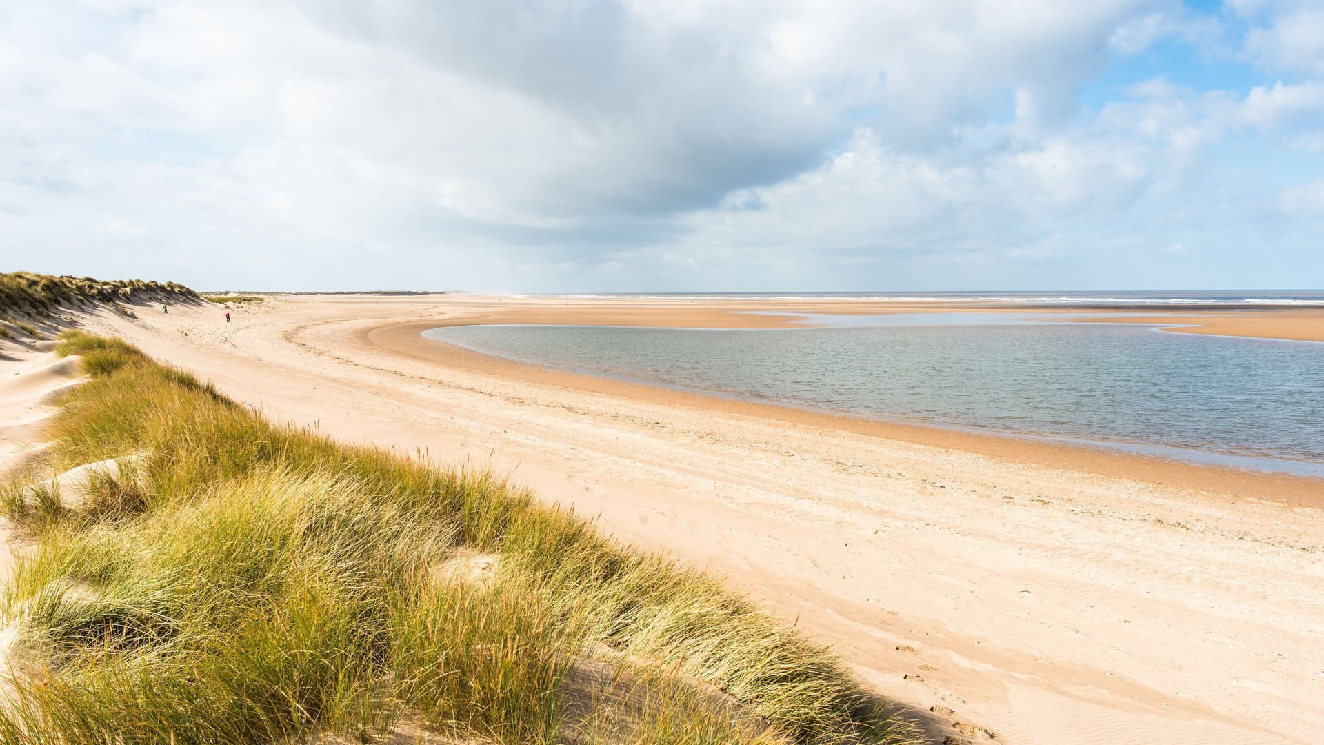 A panoramic view of the vast, sandy expanse of Holkham Beach in Norfolk, England. The image shows a serene tidal lagoon, rolling dunes covered with marram grass, and a wide blue sky with scattered clouds.