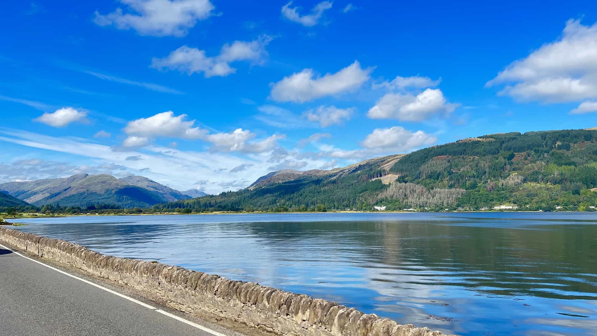 A beautiful scenic landscape of Holy Loch in Scotland, featuring calm water reflecting the blue sky and lush, green hills in the background.