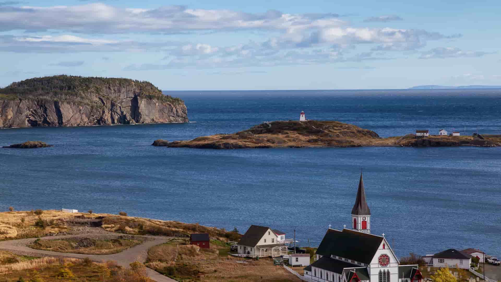 A beautiful coastal landscape of Hopedale, Newfoundland, featuring a white church with a steeple in the foreground and a lighthouse on a small island in the distance.