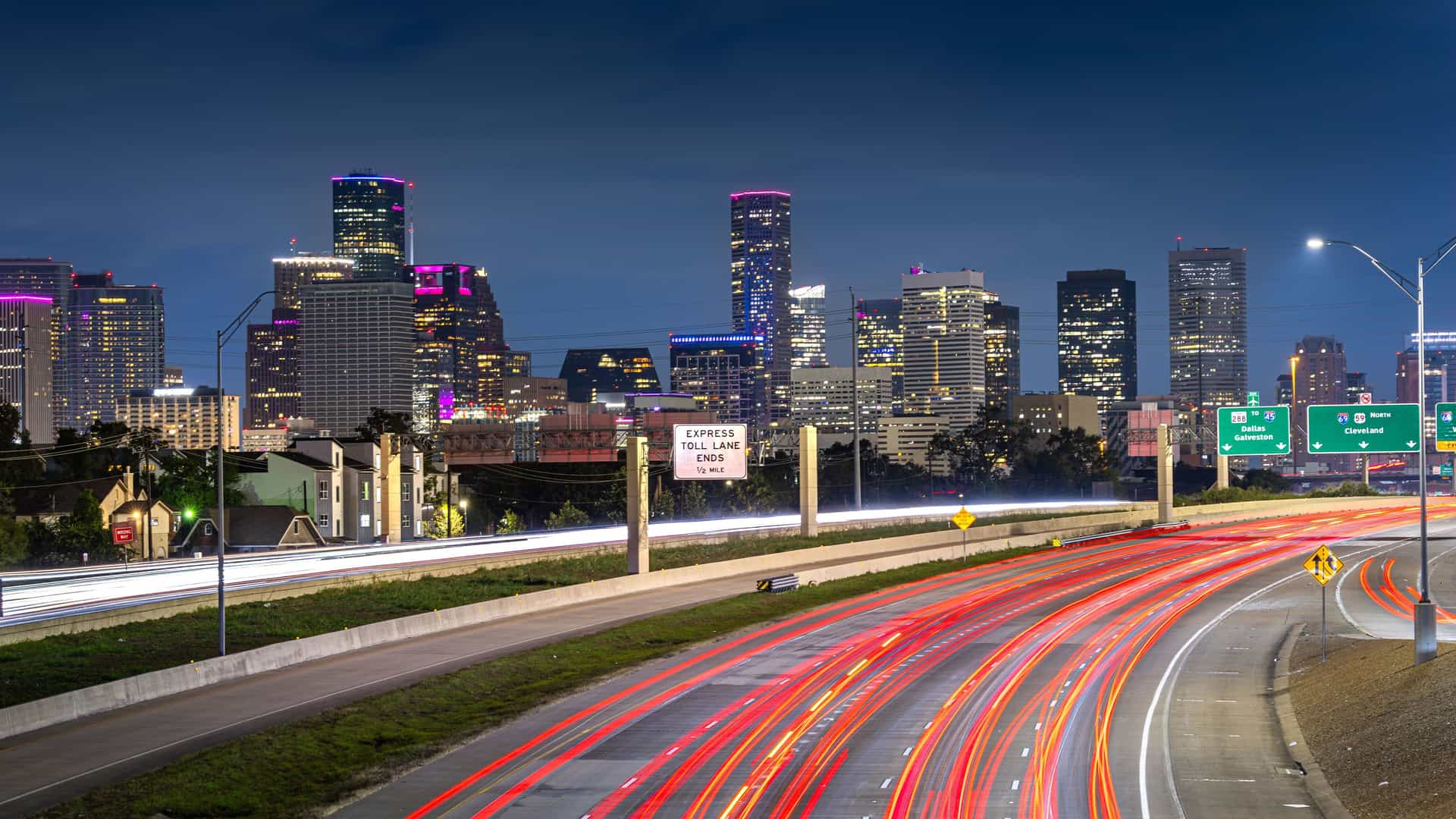 "A long-exposure, nighttime shot of the vibrant Houston, Texas, skyline, with the downtown skyscrapers illuminated against a blue twilight sky and the red light trails from cars on the busy highway in the foreground. "