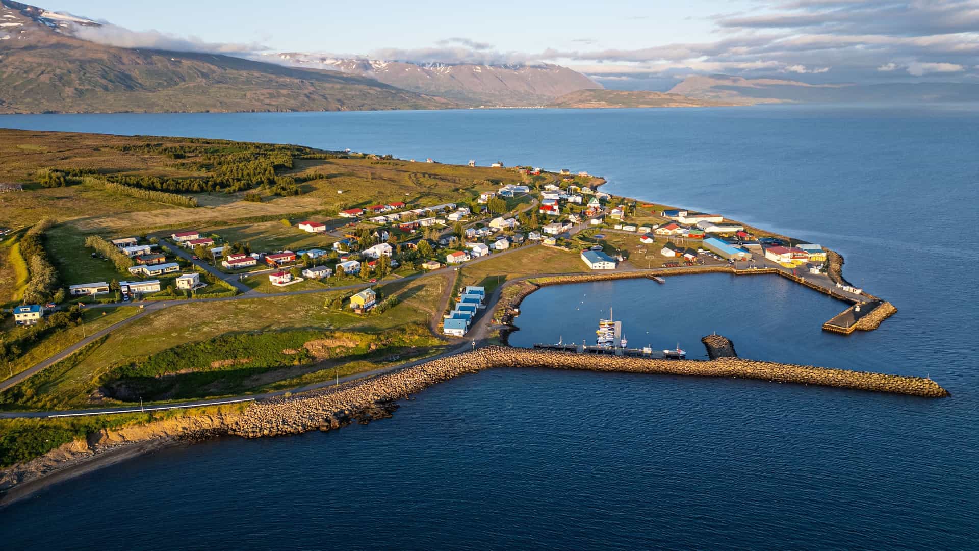 An aerial view of Hrisey, Iceland, showing the small fishing village with colorful houses, a harbor, and a stunning backdrop of the fjord and mountains.