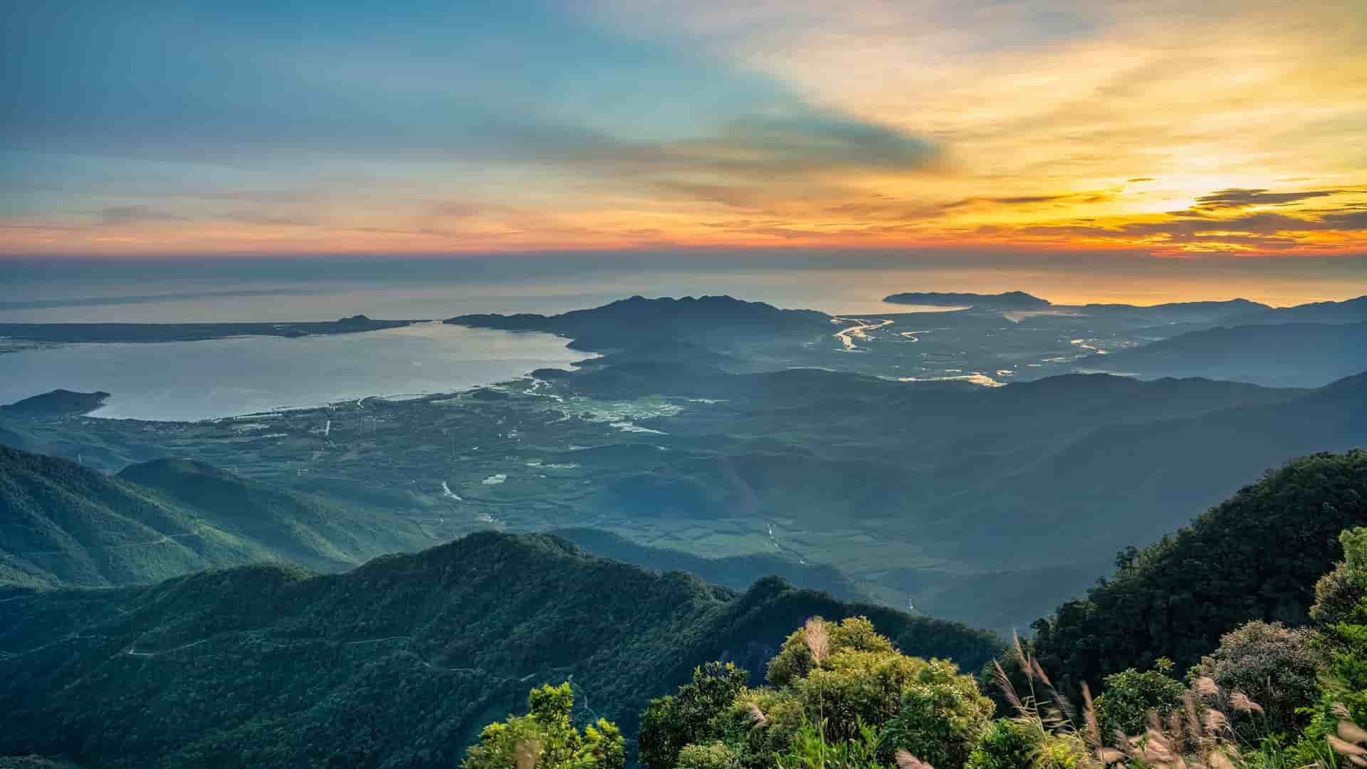A dramatic aerial shot of the Hai Van Pass near Hue, Vietnam, with a bay and coastline visible between lush green mountains under a vibrant sunrise sky.